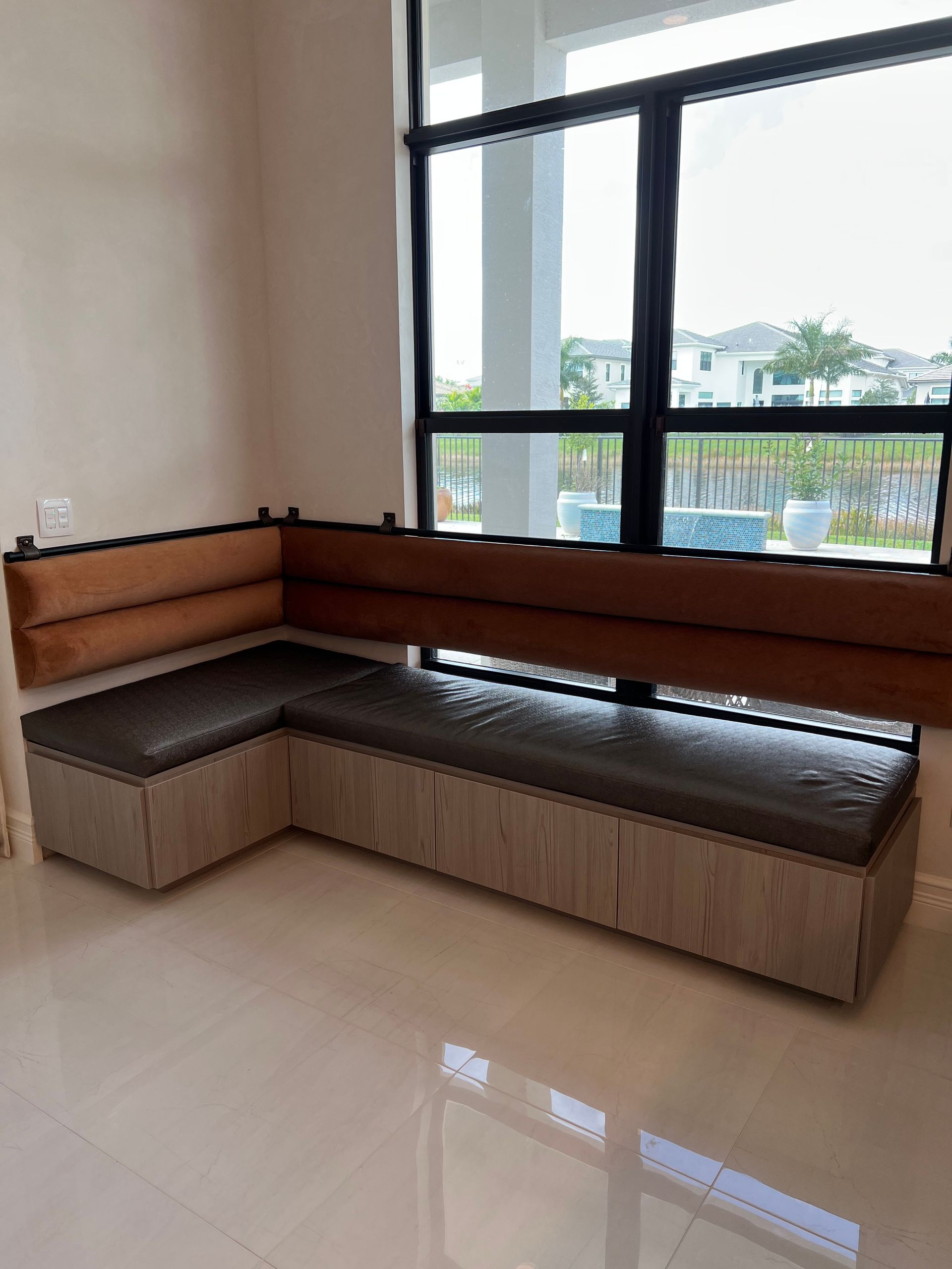 An L-shaped custom built-in wooden window bench with dark gray cushioned seats and brown padded backrests.