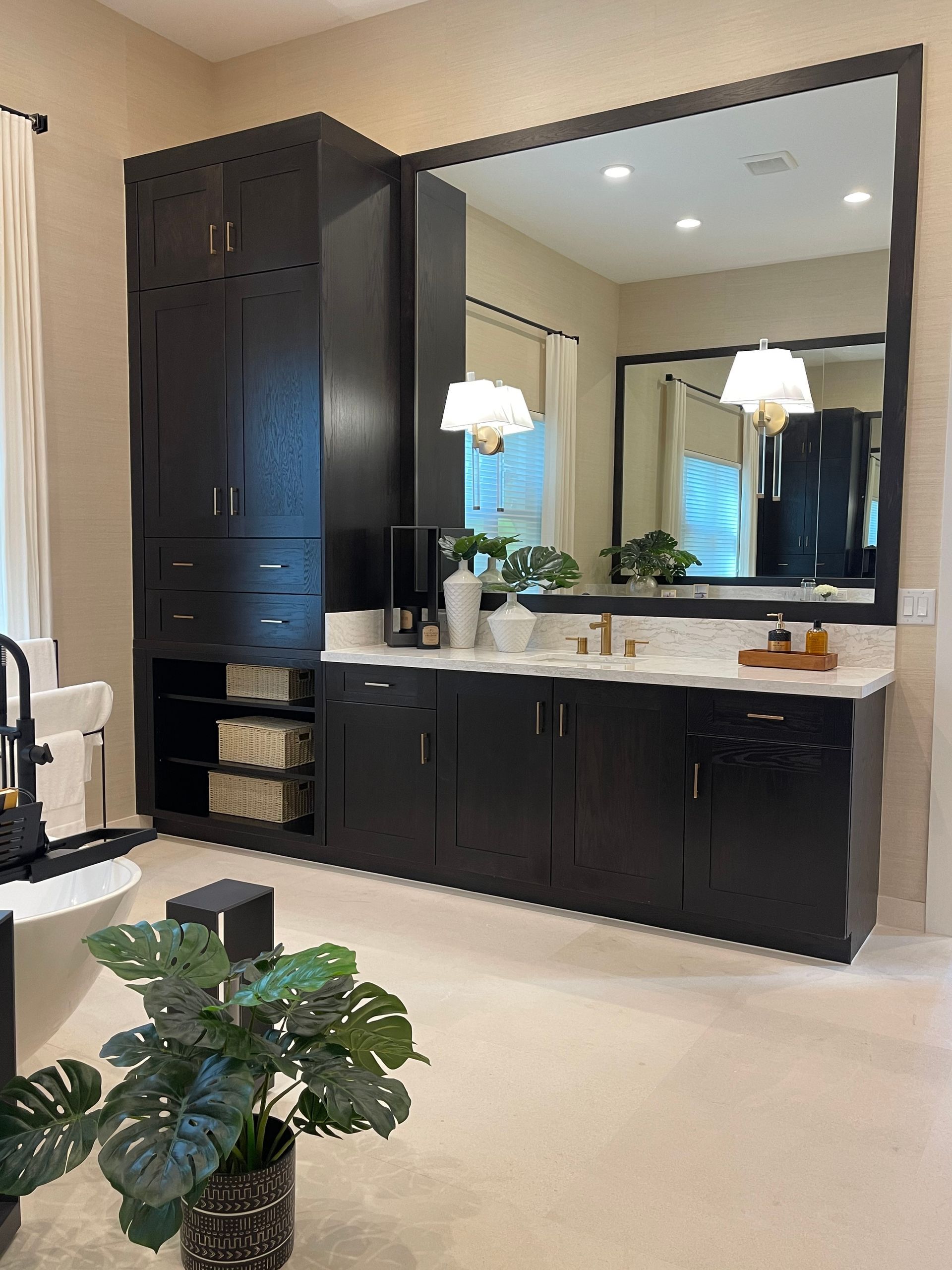 A modern bathroom with dark wood cabinets, a large mirror, white countertops, and a potted monstera plant in the foreground.