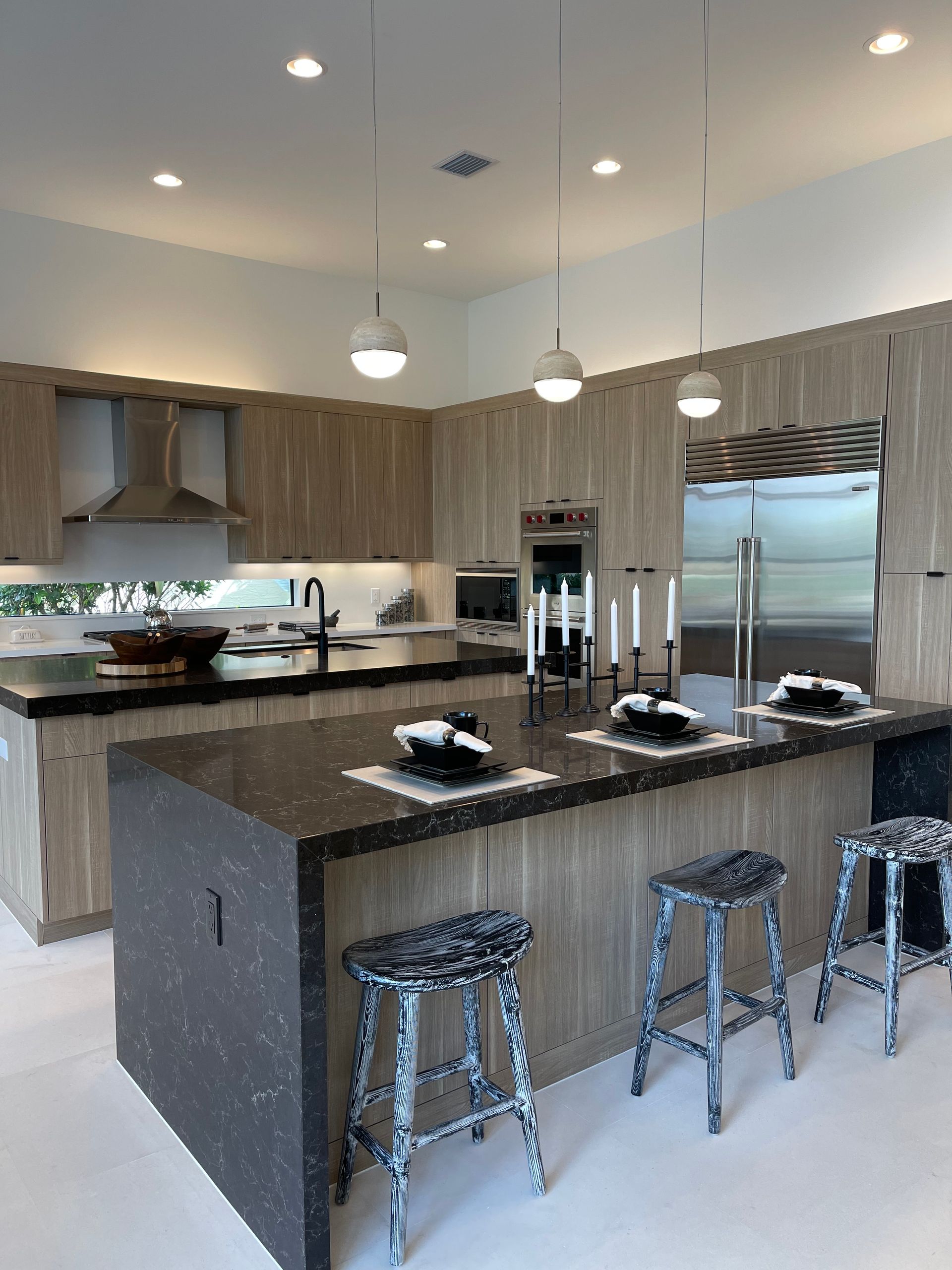 A modern kitchen with light wood cabinets, dark speckled countertops, three pendant lights, and matching bar stools.