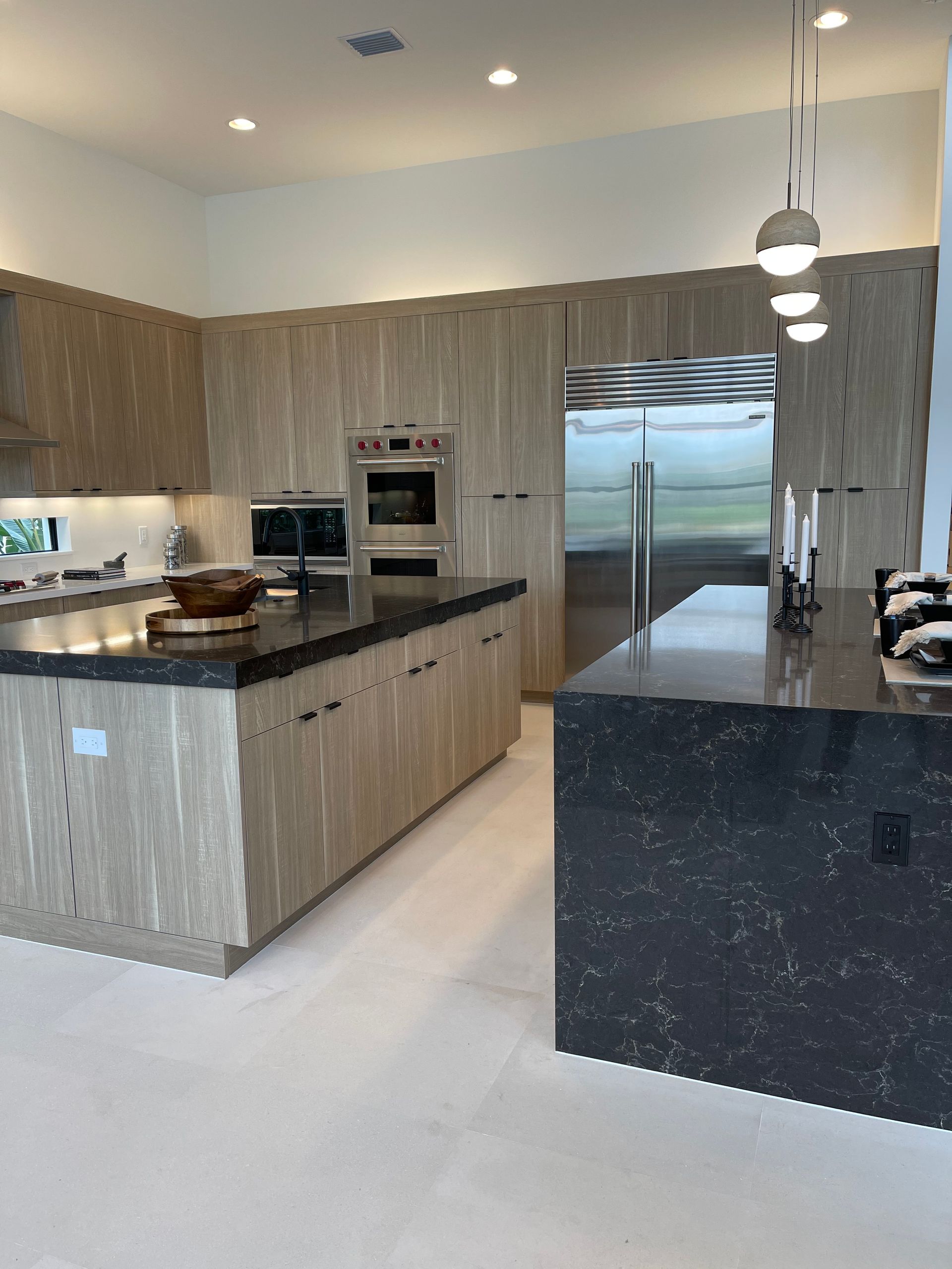 A modern kitchen featuring light wood-grain cabinetry, black countertops on two islands, and a stainless steel refrigerator.