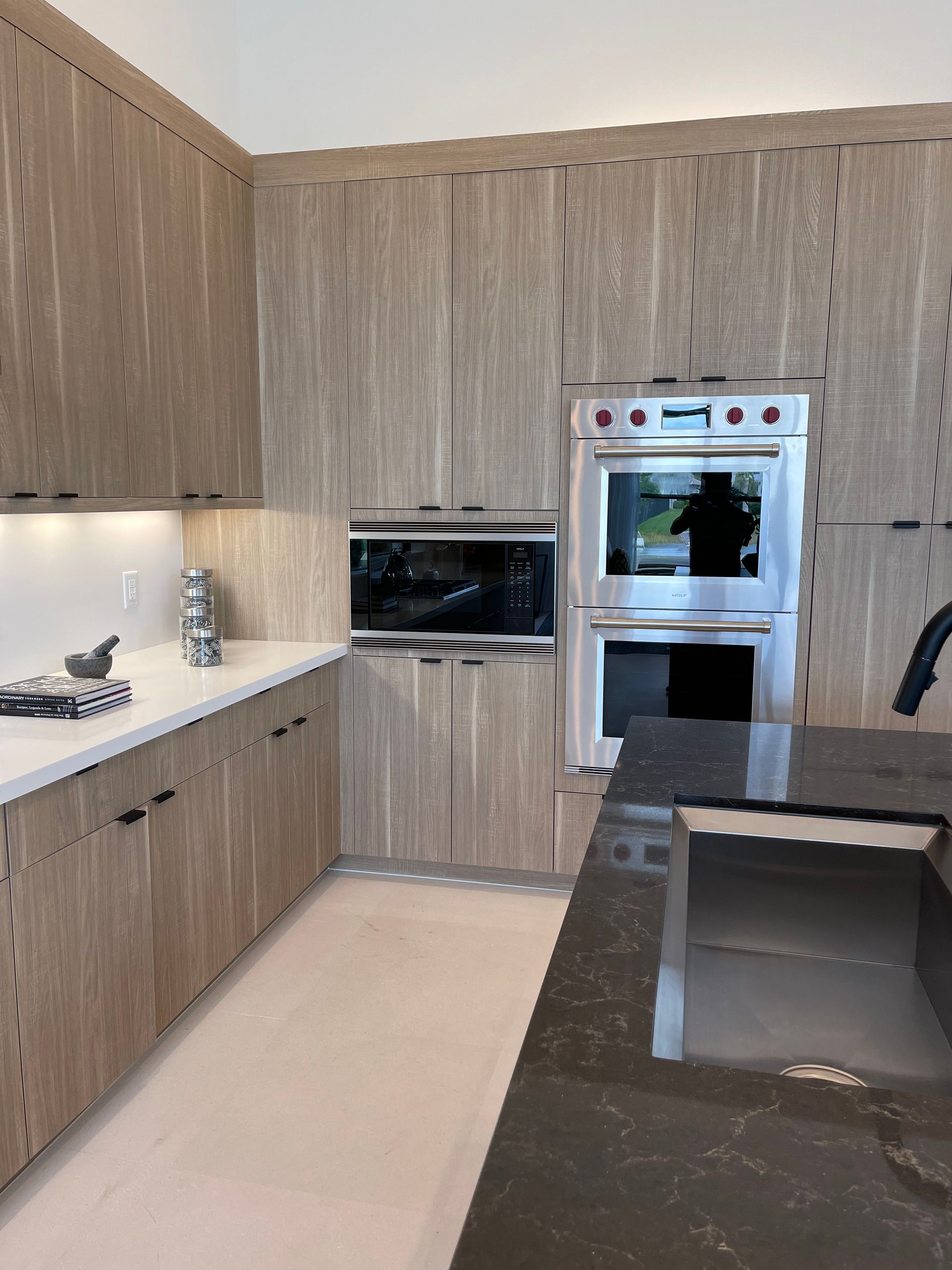 A modern kitchen with light wood cabinetry, white countertops, stainless steel double ovens, and a dark stone island.