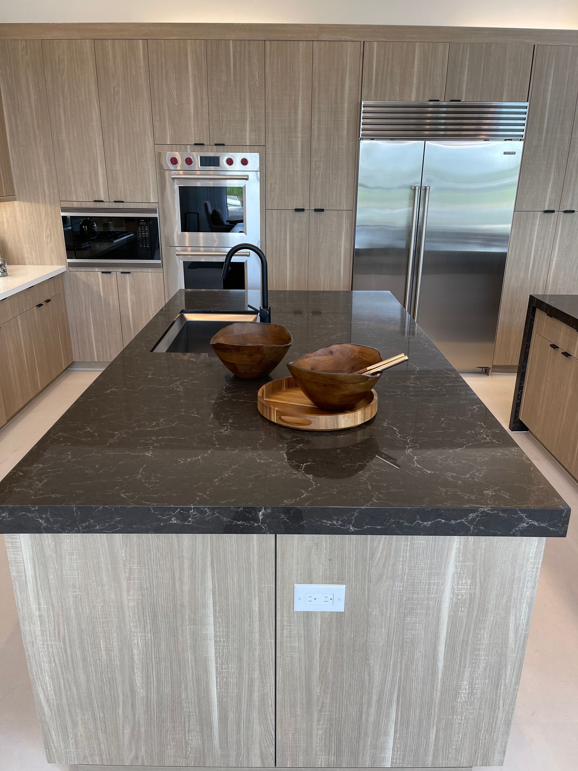 A modern kitchen with a large dark countertop island, light-wood cabinets, stainless steel appliances, and wood bowls.