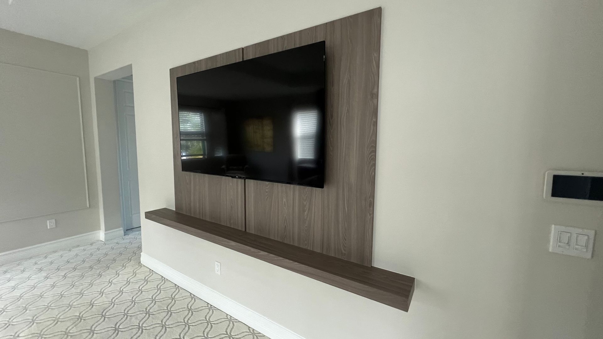 A mounted television on a wooden accent wall panel above a floating shelf in a modern, neutral-toned room.