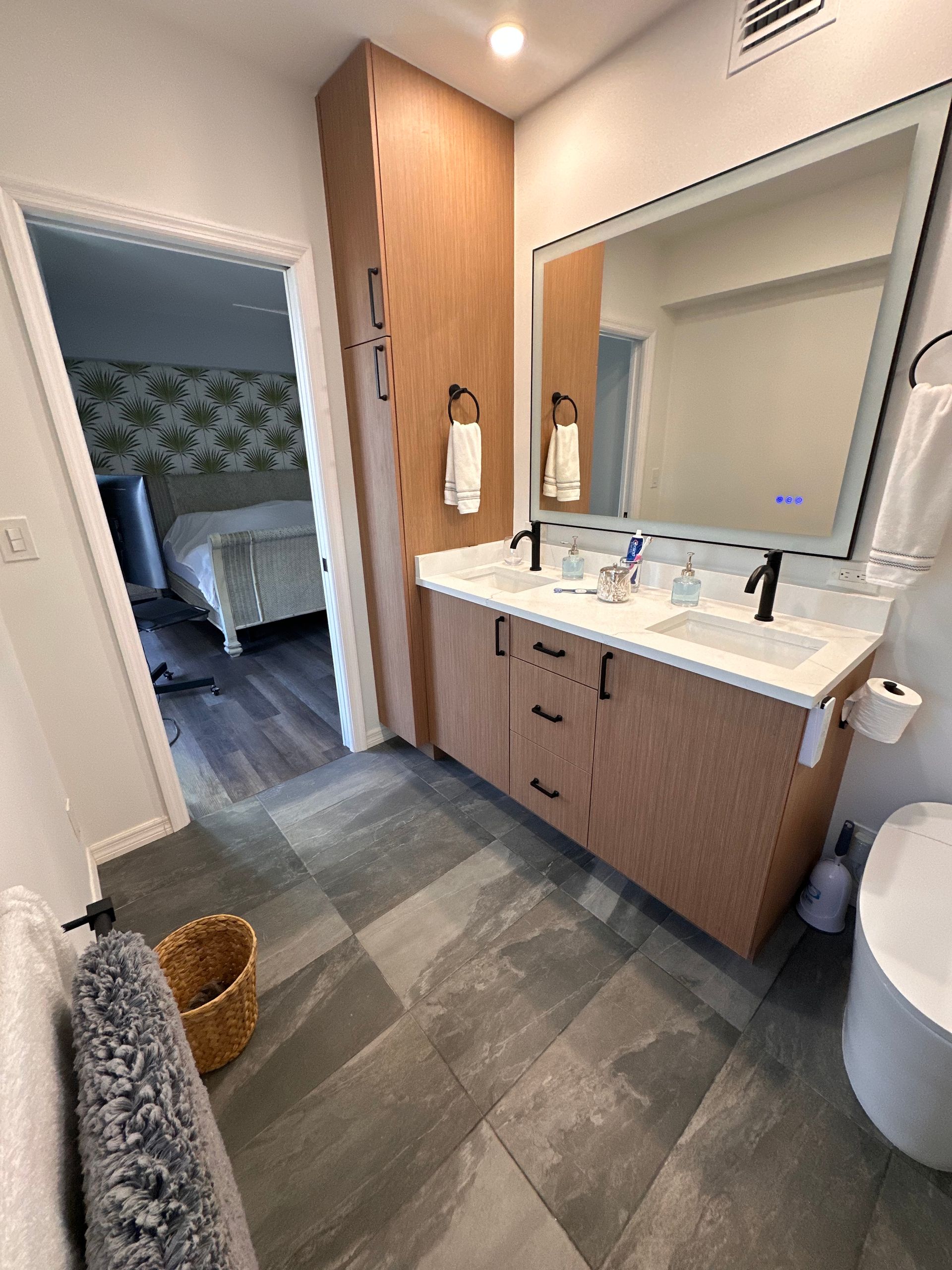 Modern bathroom with light wood cabinets, double vanity, grey stone tile floor, and a view into a bedroom.