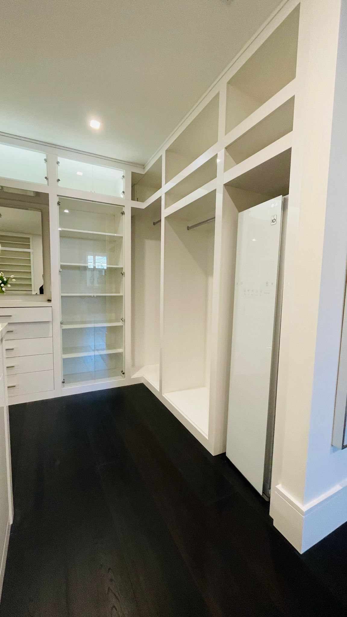 An empty walk-in closet with white shelving, hanging rods, and a tall mirror against a white wall on dark flooring.