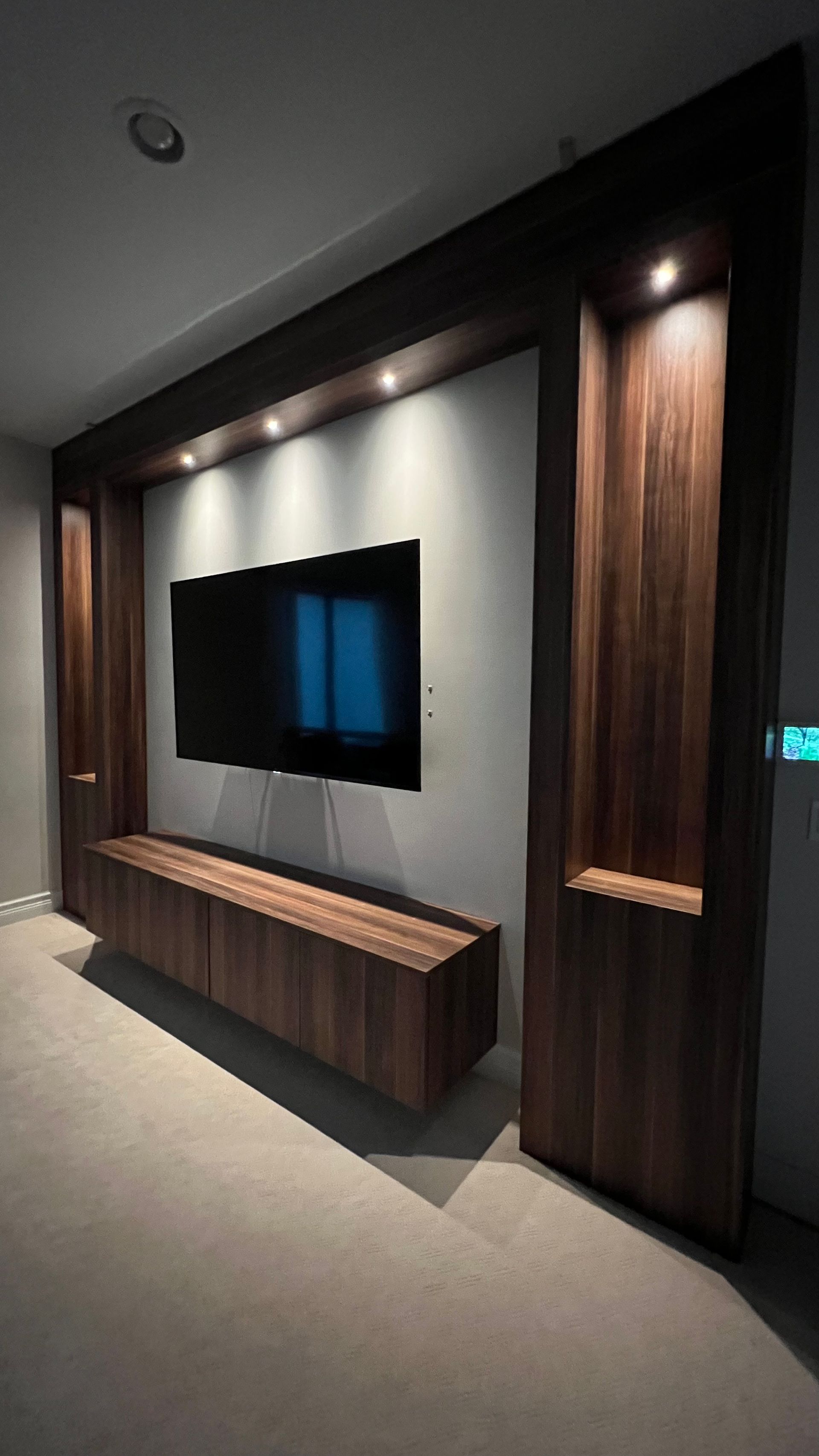 Wall-mounted television centered within a dark wood frame featuring integrated shelves and recessed ceiling lighting.