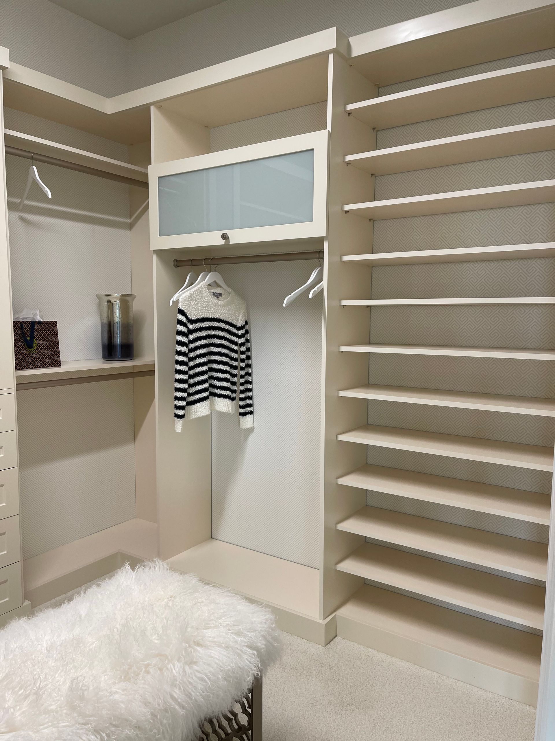 A beige walk-in closet featuring shelving, a hanging rack with a striped sweater, a frosted cabinet, and a furry stool.