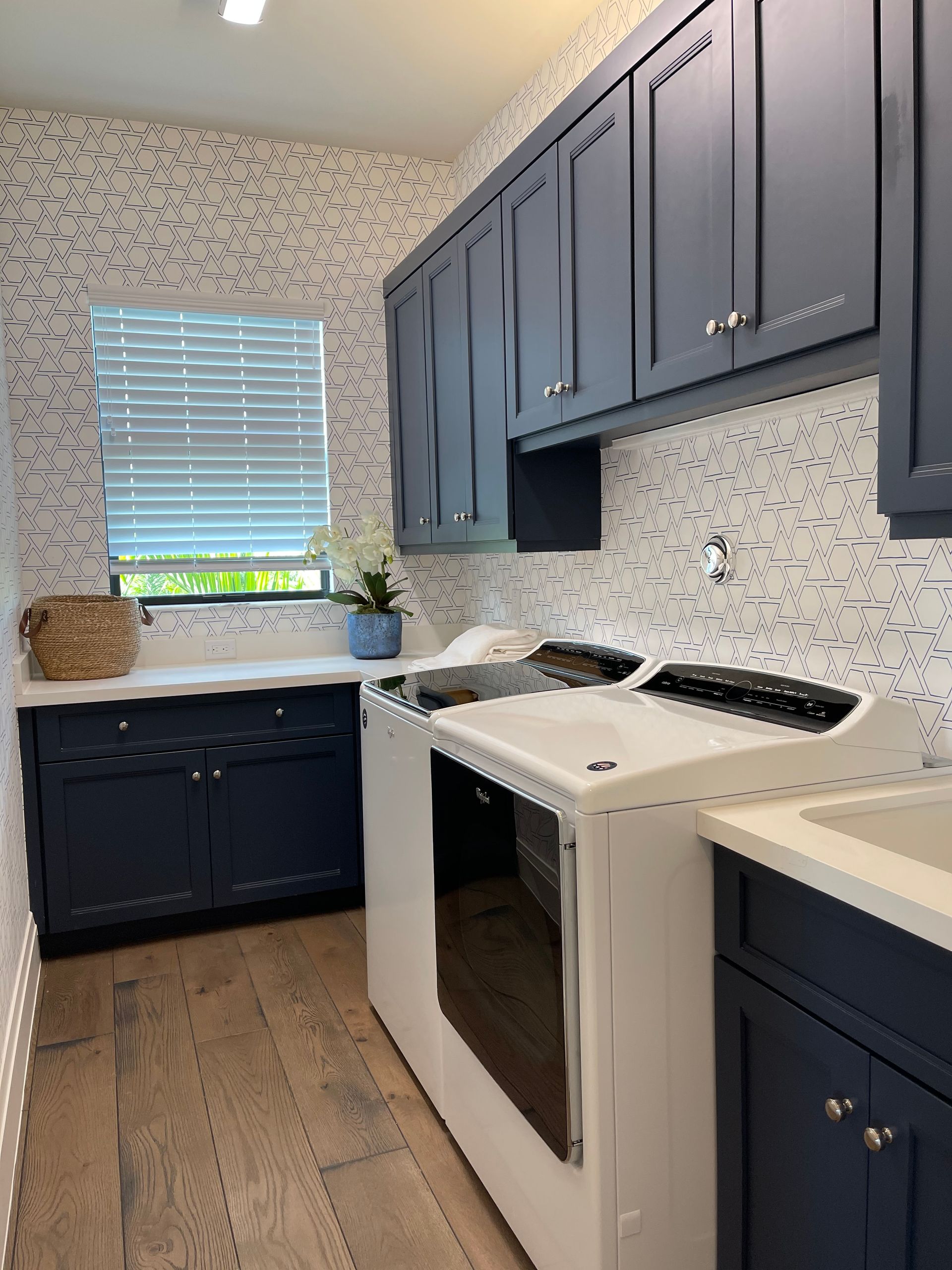 A laundry room with dark blue cabinets, white patterned wallpaper, a white appliance, and light wood flooring.