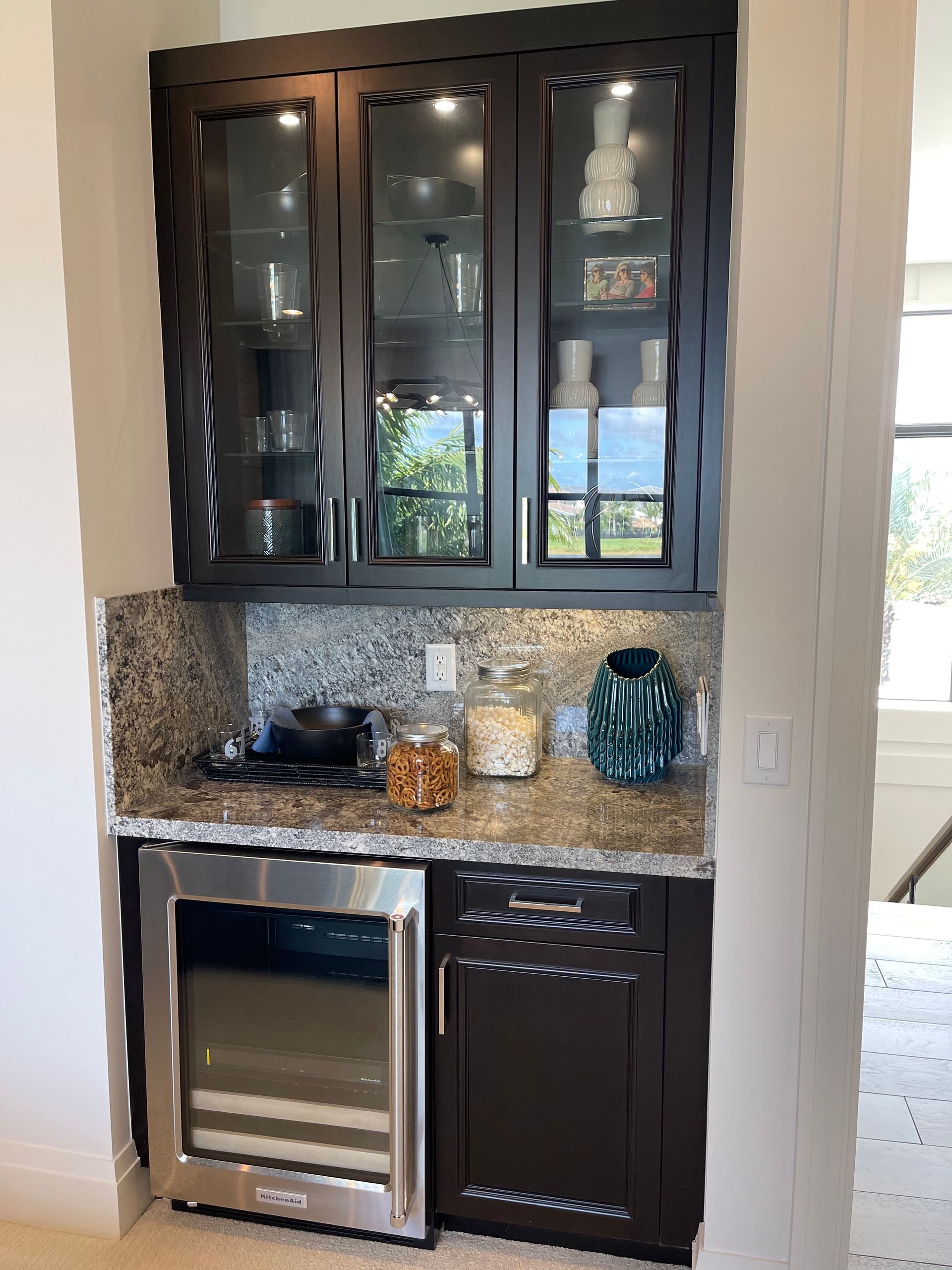 A built-in dark wood wet bar featuring glass-front upper cabinets, granite countertops, and a stainless steel wine cooler.