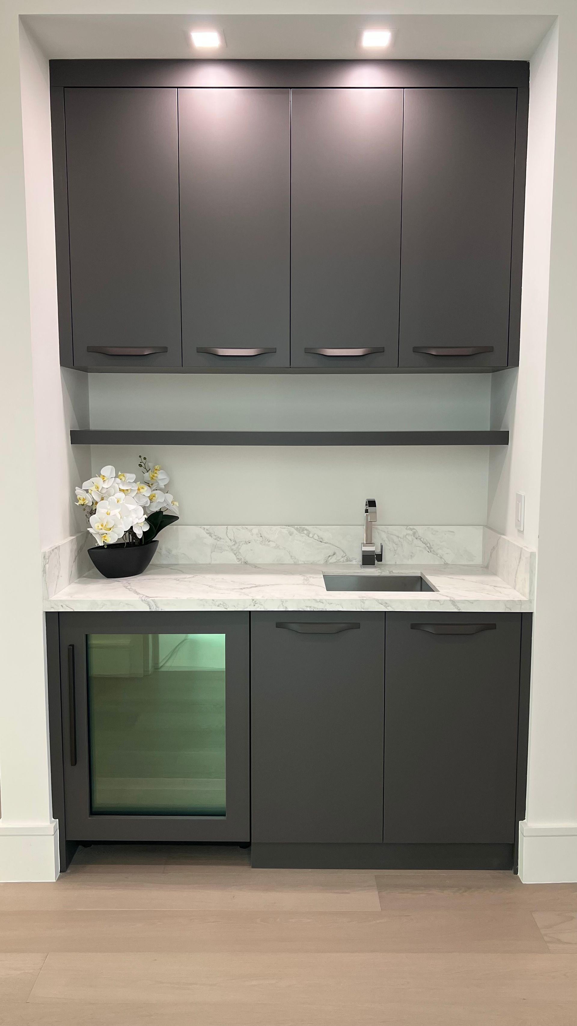 A modern wet bar featuring dark gray cabinetry, a marble countertop, a small sink, a wine fridge, and open shelving.