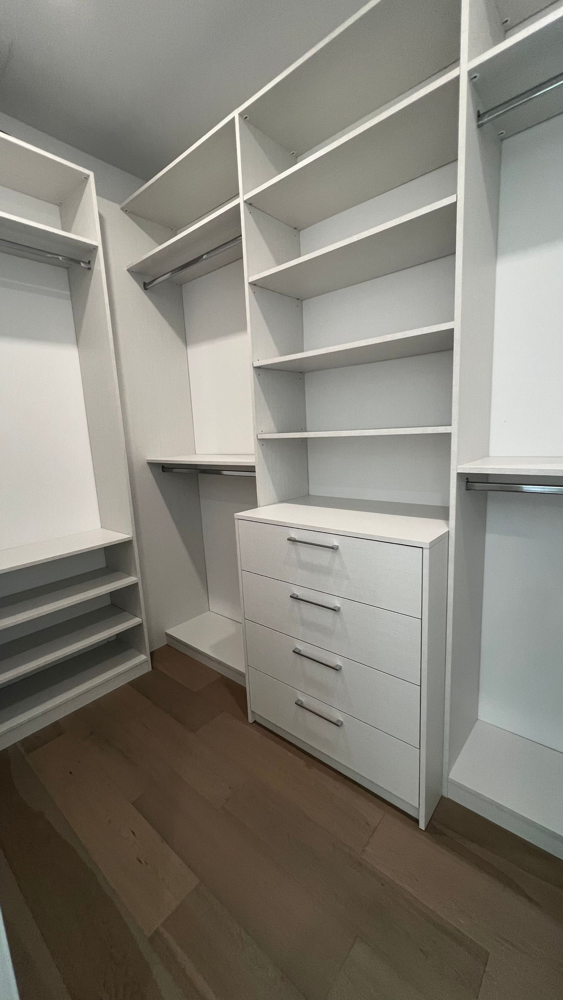 A walk-in closet featuring white wooden shelving, hanging rods, and a four-drawer dresser on light brown wood flooring.