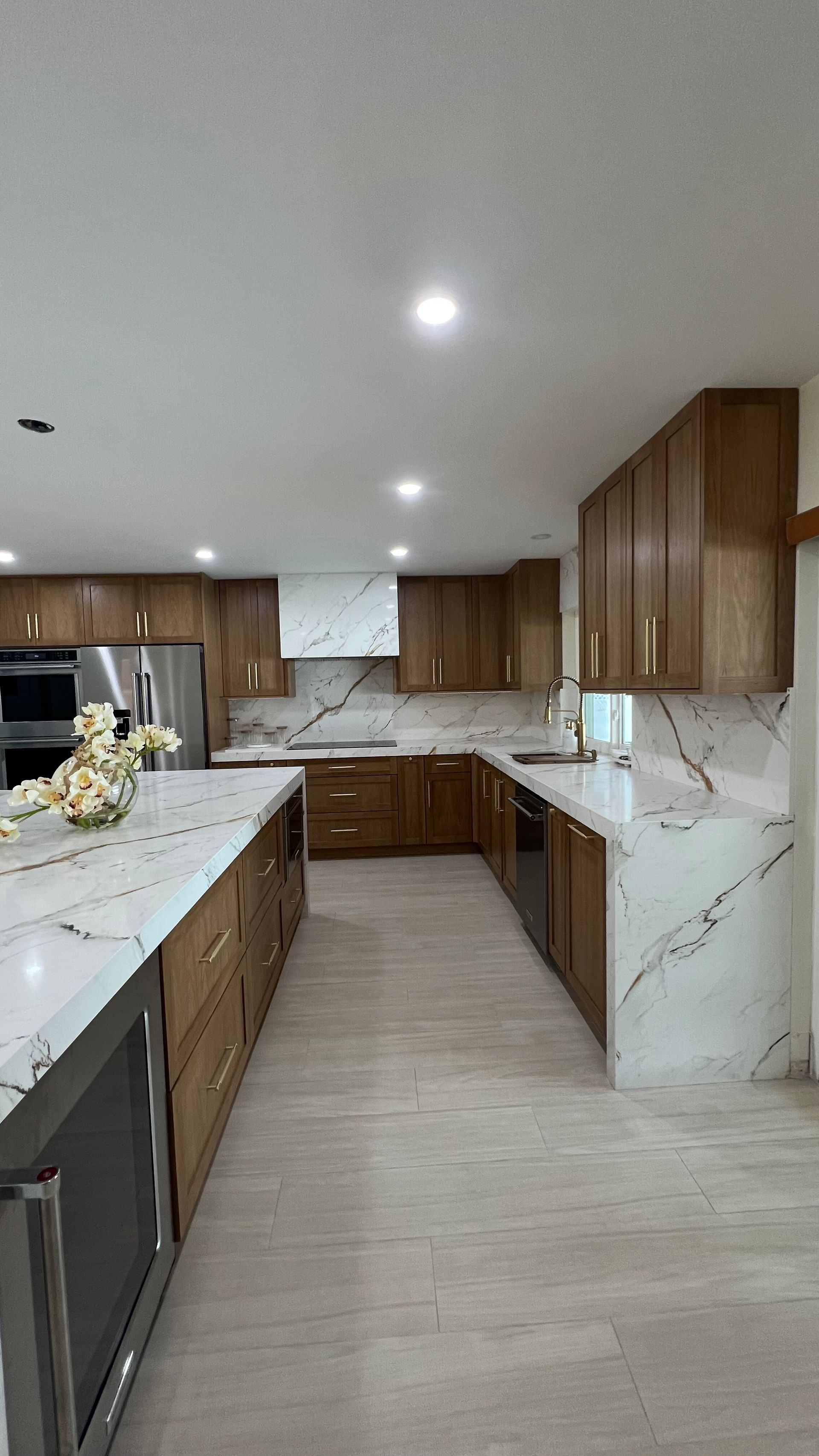 A modern kitchen featuring wooden cabinets, white marble-veined countertops, stainless steel appliances, and tile floors.