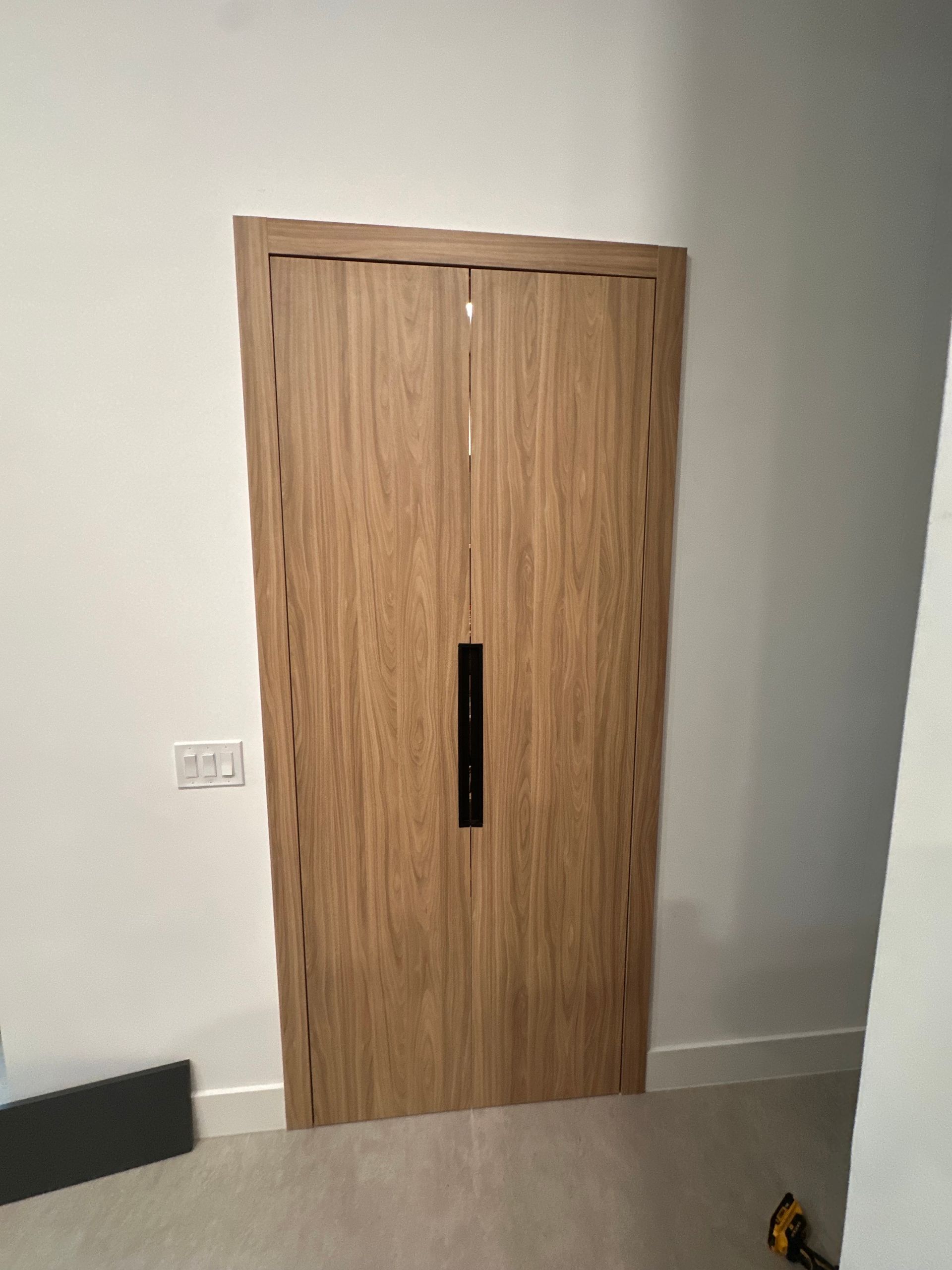 Light wood double doors with a black vertical handle set in a white wall.