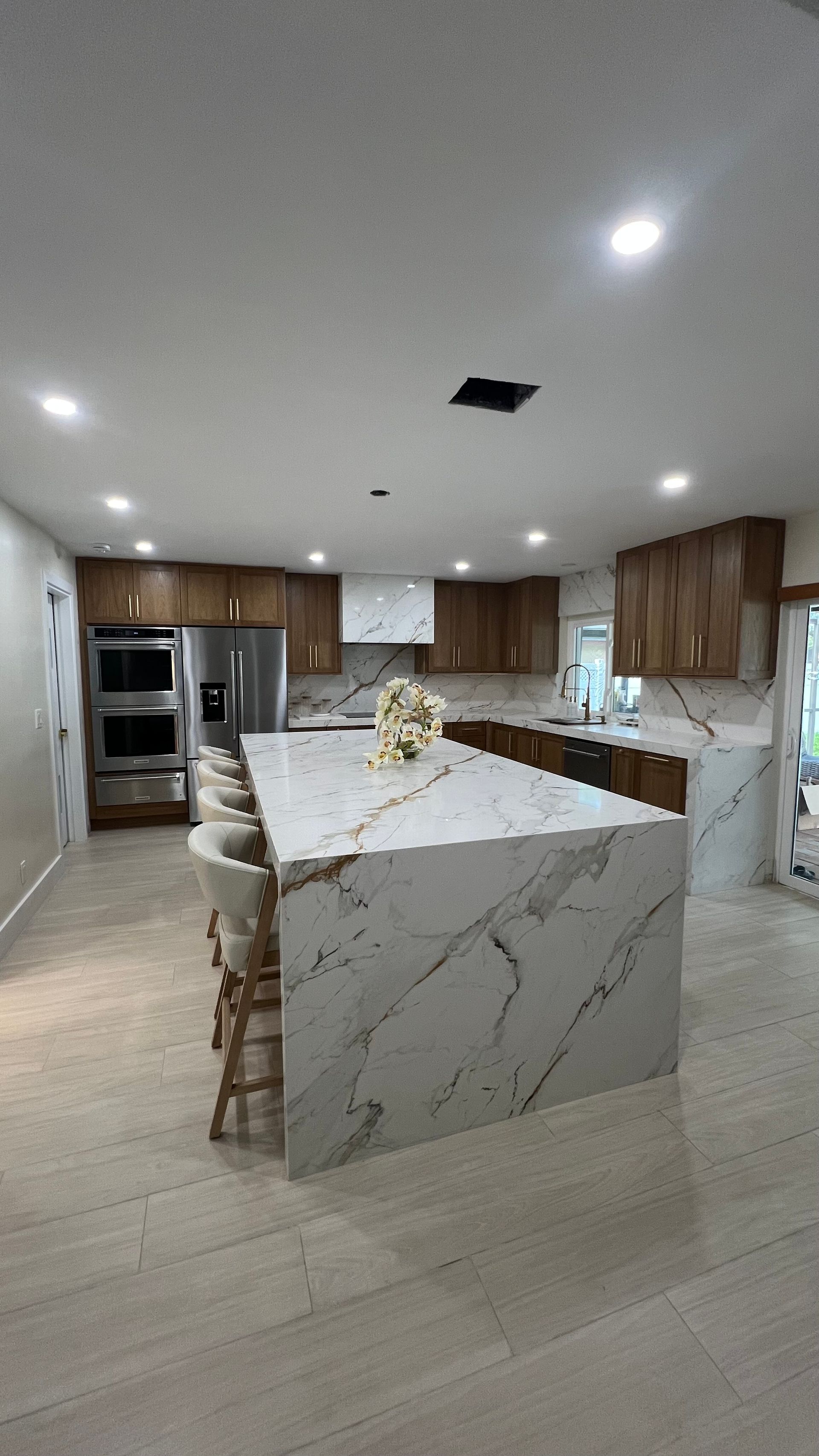 A modern kitchen features a large, marble-topped island, wooden cabinetry, stainless steel appliances, and tile flooring.