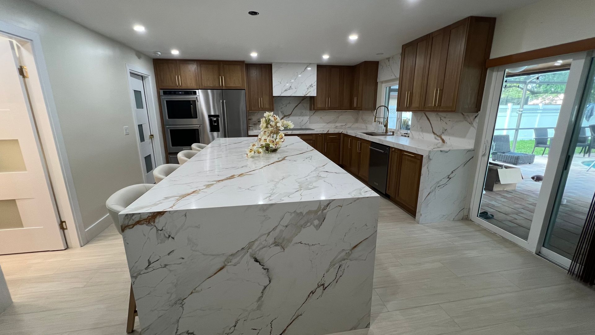A modern kitchen with a large marble waterfall island, wood cabinets, stainless steel appliances, and tile flooring.