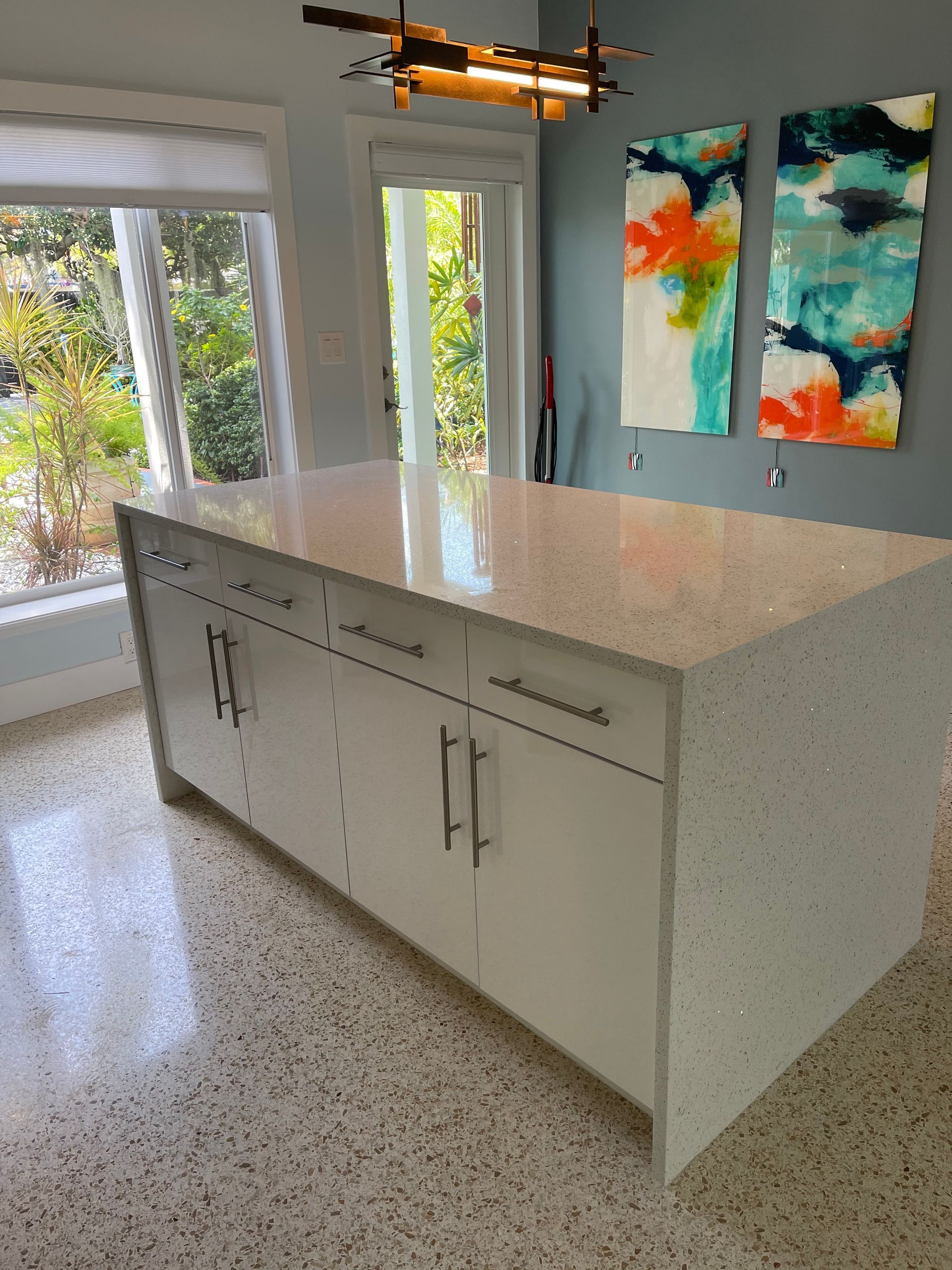 A white kitchen island with a speckled stone countertop, set in a room with terrazzo floors and abstract wall art.