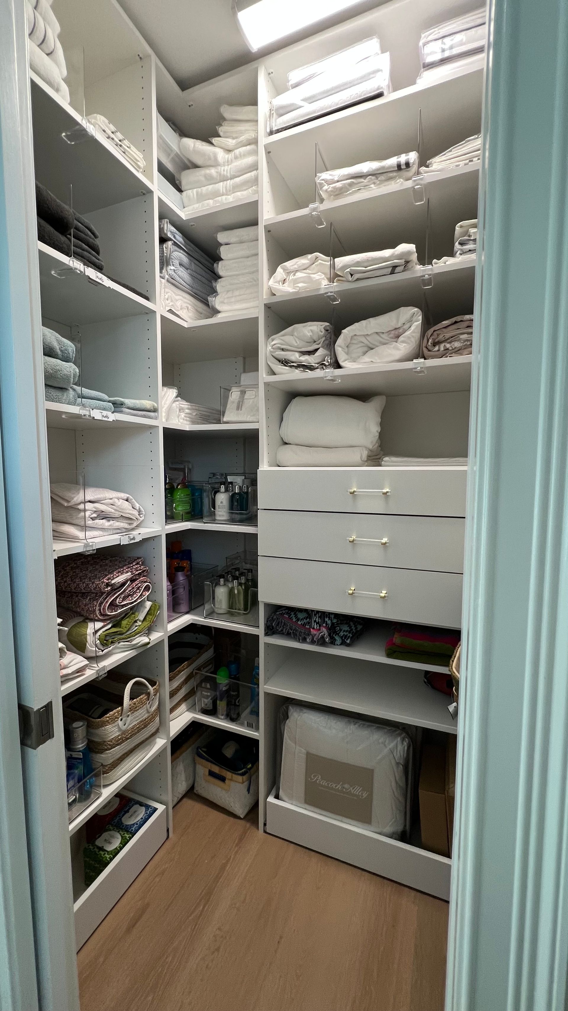 A walk-in linen closet with organized white shelves, stacked towels, blankets, linens, and three drawers on light wood floors.