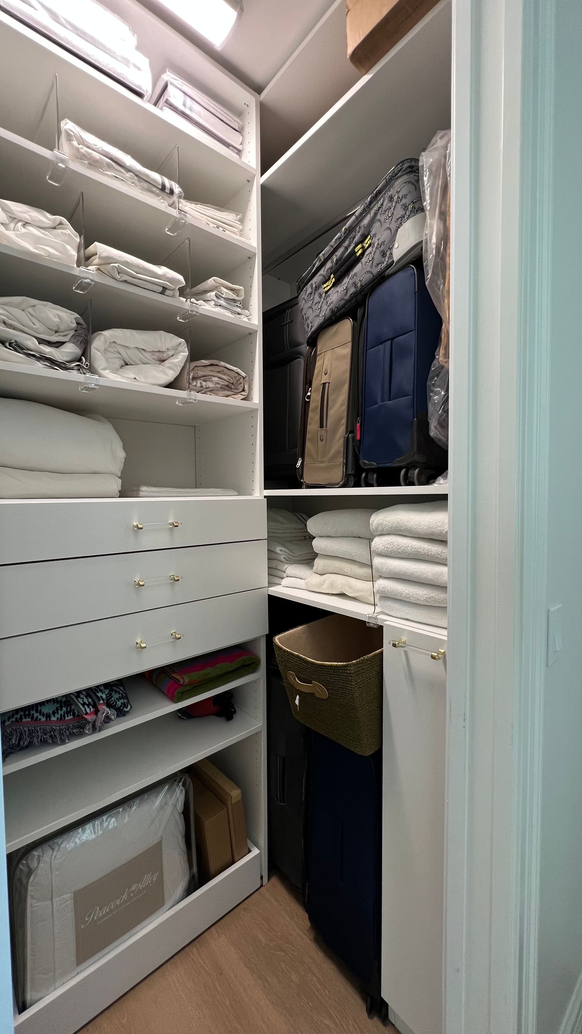 A white closet with shelves filled with folded linens, drawers, and upright suitcases stored on the side.
