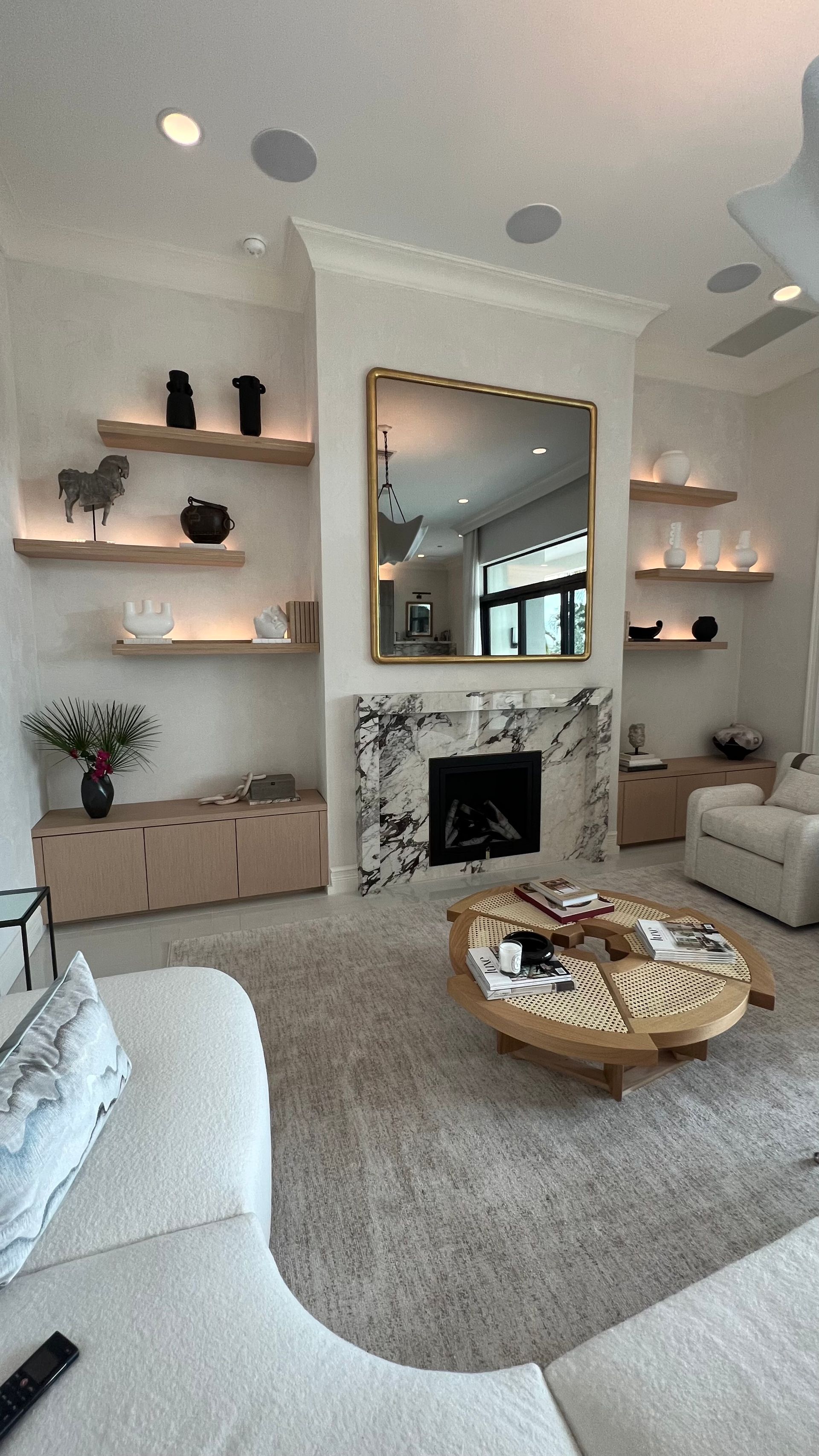 A bright living room featuring a marble fireplace, floating shelves with decor, a round coffee table, and a sectional.