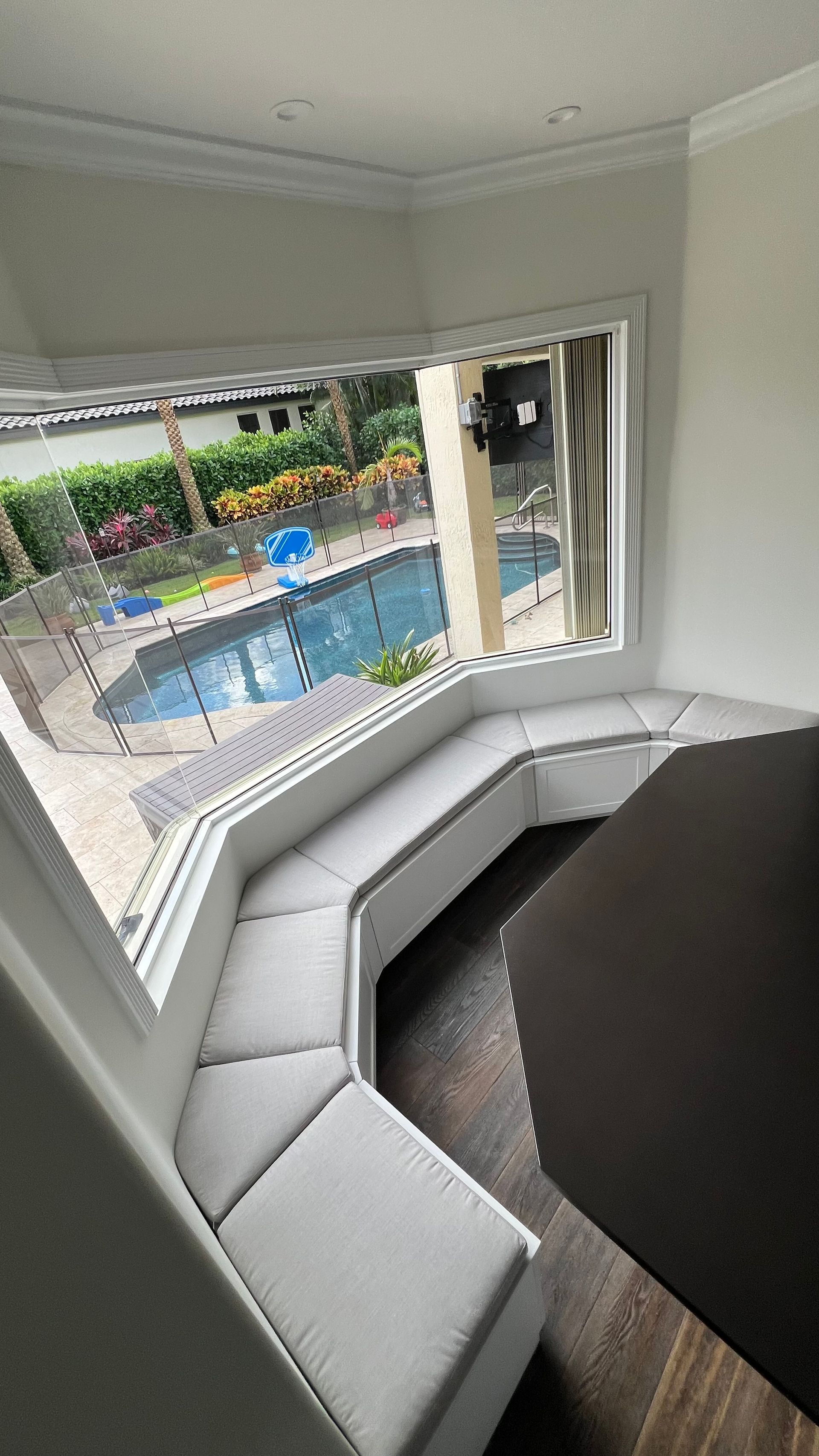 A bright, curved built-in window bench with grey cushions overlooks a backyard swimming pool and lush landscaping.