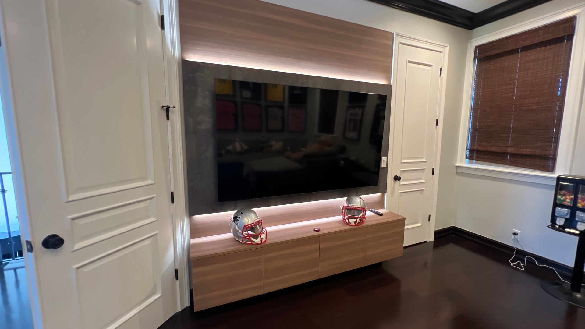 A modern entertainment wall featuring a mounted television, LED backlighting, and a wooden console with two football helmets.