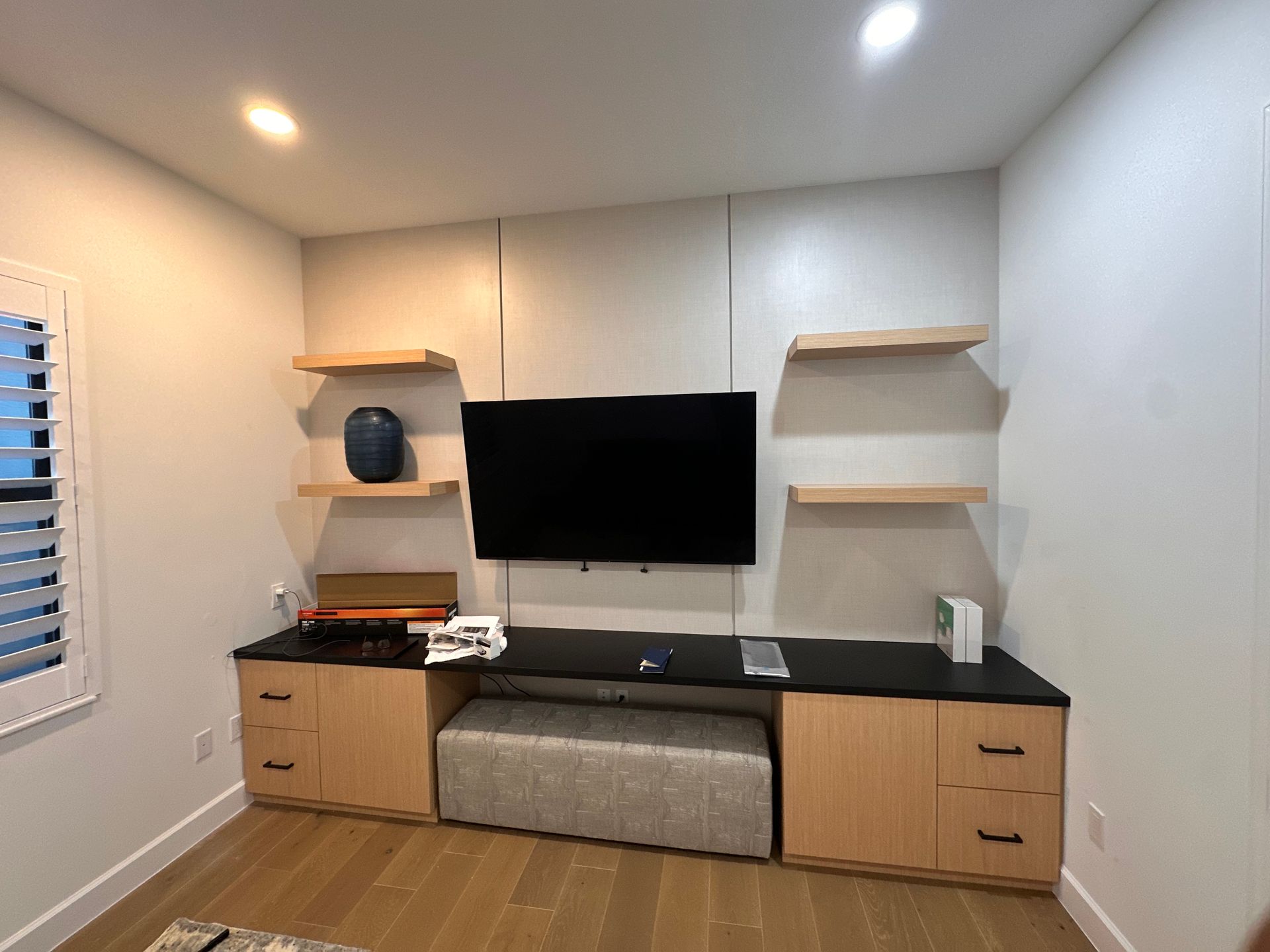 A wall-mounted TV flanked by floating wooden shelves above light wood cabinets with a grey upholstered bench underneath.