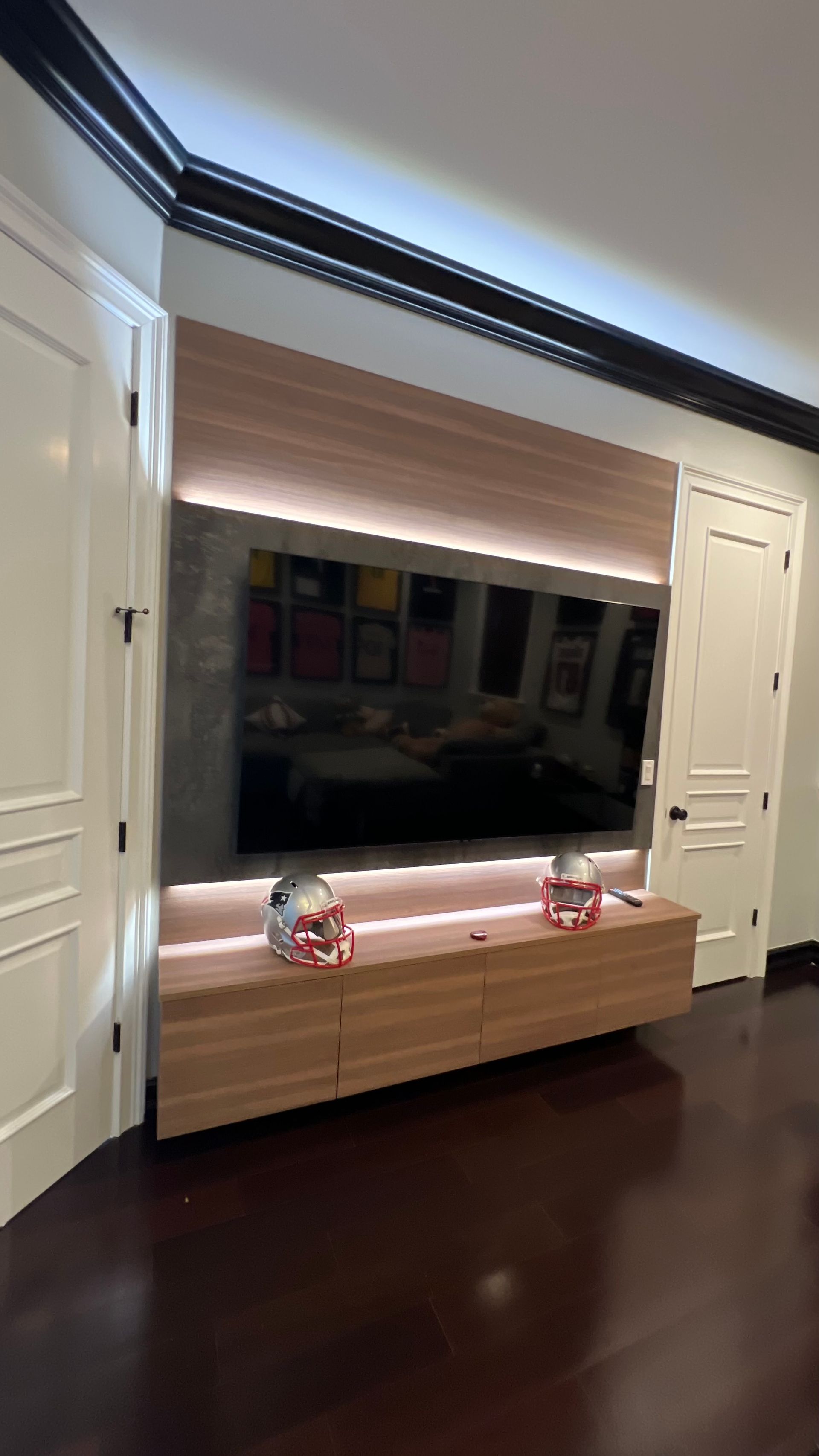 A wall-mounted TV framed by wood paneling and LED lighting, centered between two white doors above a matching console.