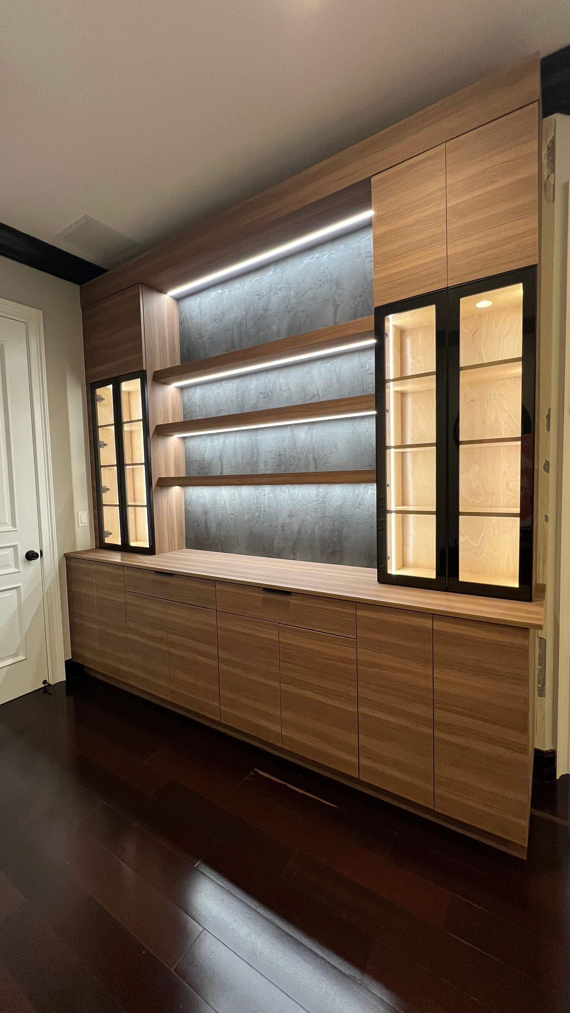 Custom wood wall unit featuring lower cabinets, open shelving with under-cabinet lighting, and glass-front display cases.