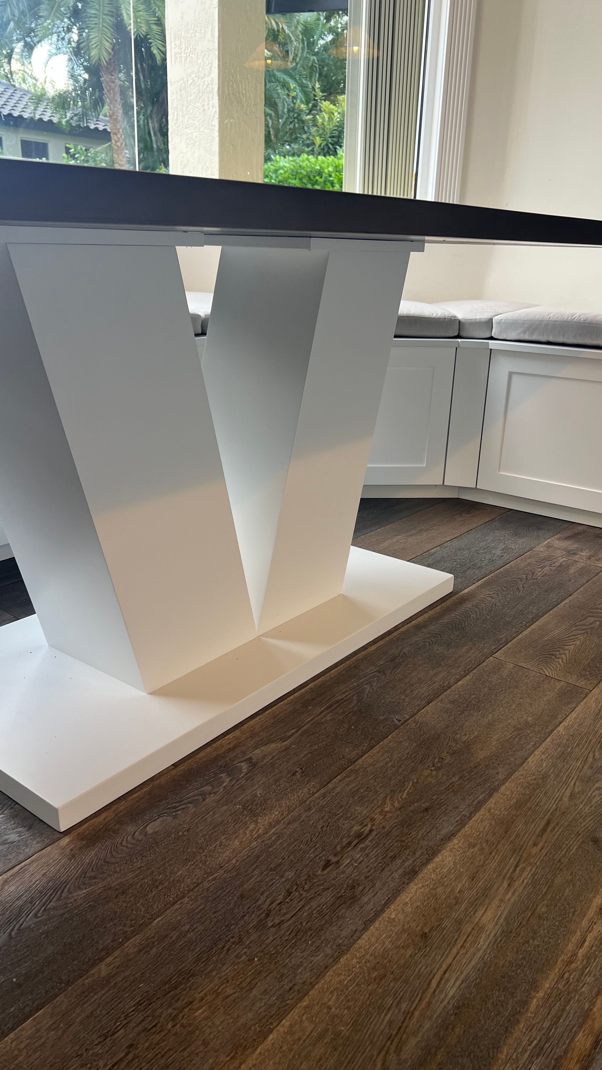 A white V-shaped table base on a dark wooden floor, positioned near white built-in bench seating with gray cushions.