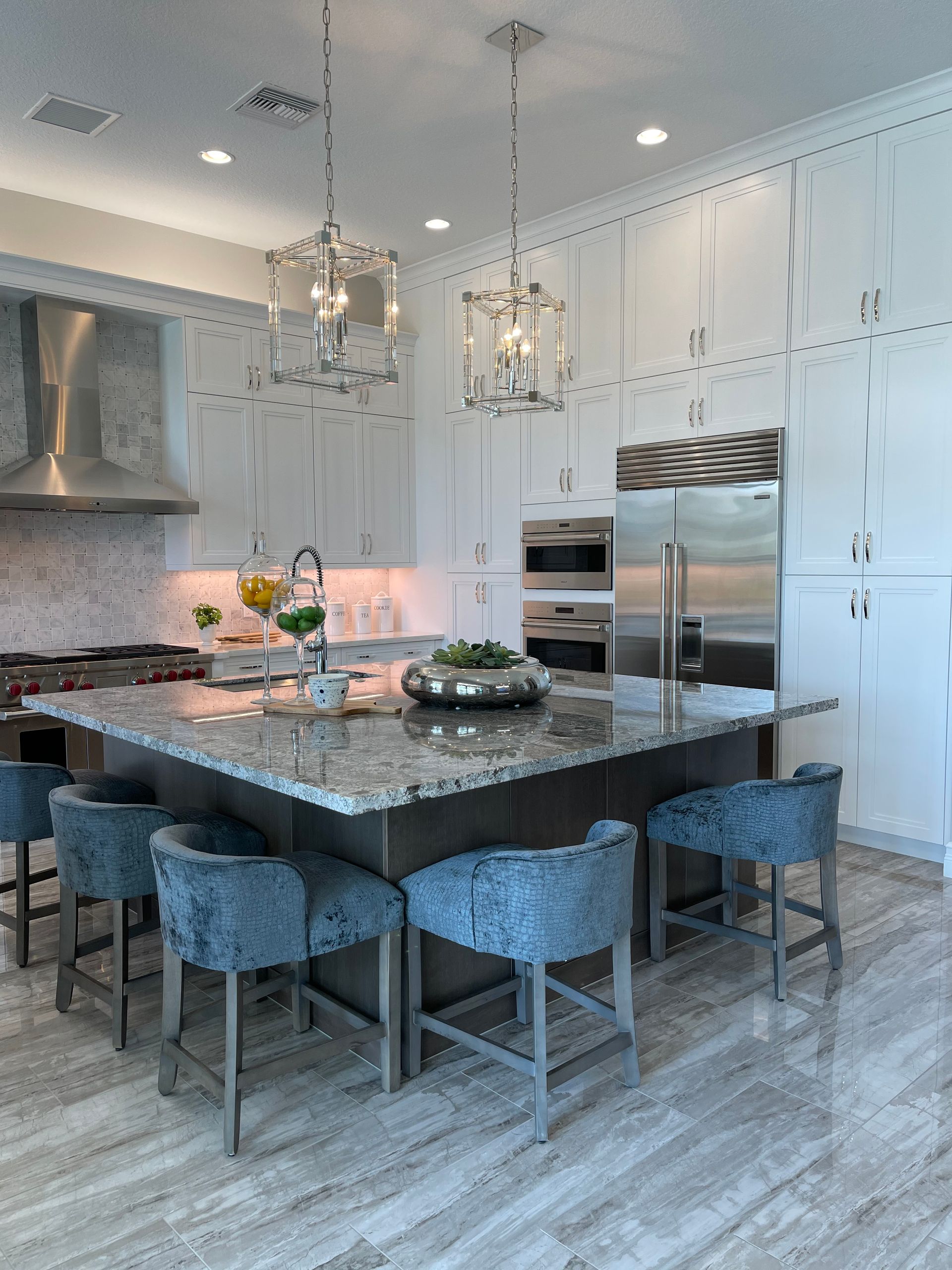 A modern kitchen with a large granite island, blue upholstered bar stools, white cabinetry, and stainless steel appliances.