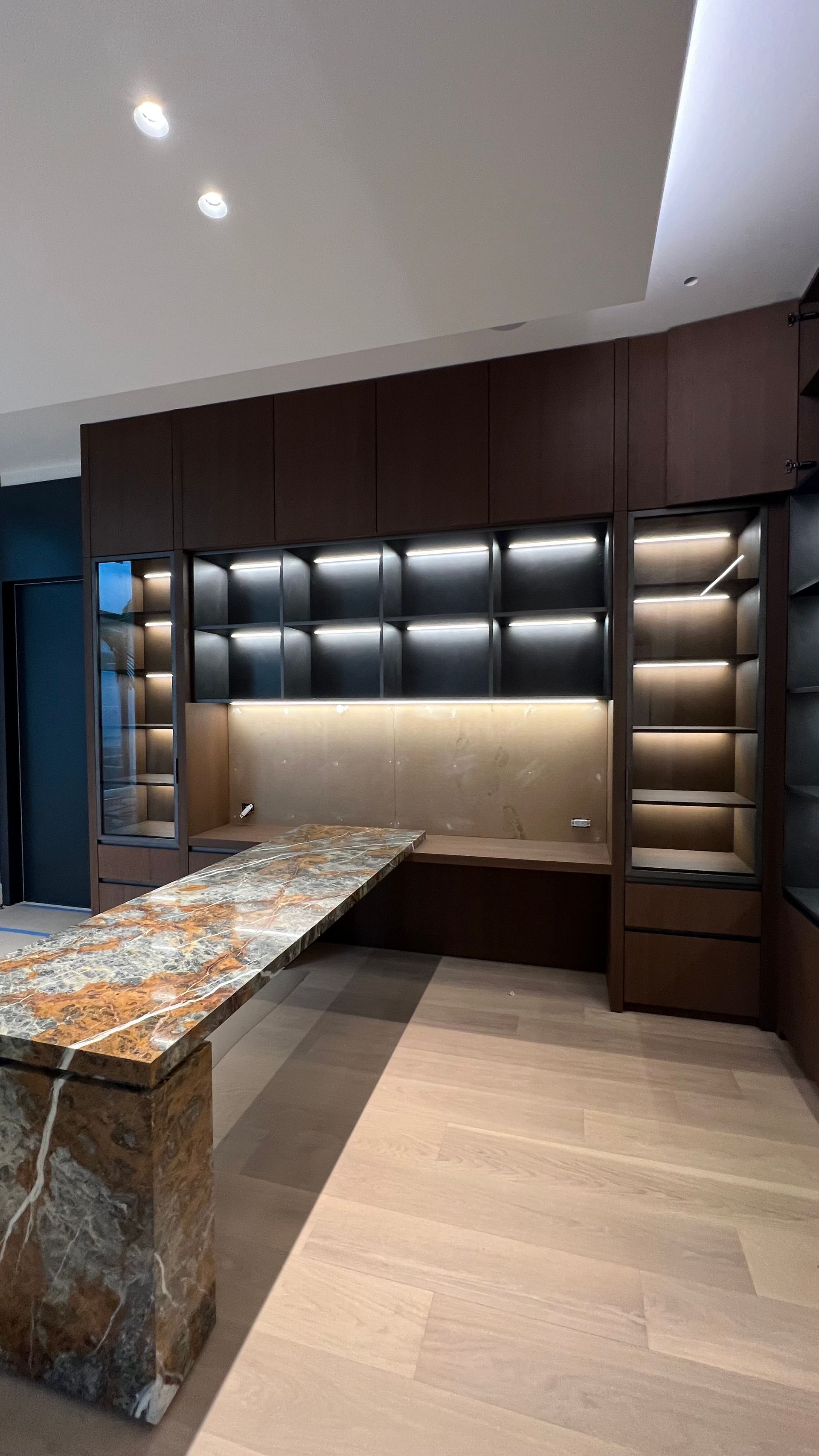 Modern home office with a dark wood wall unit, illuminated glass shelving, and a large, patterned stone desk island.