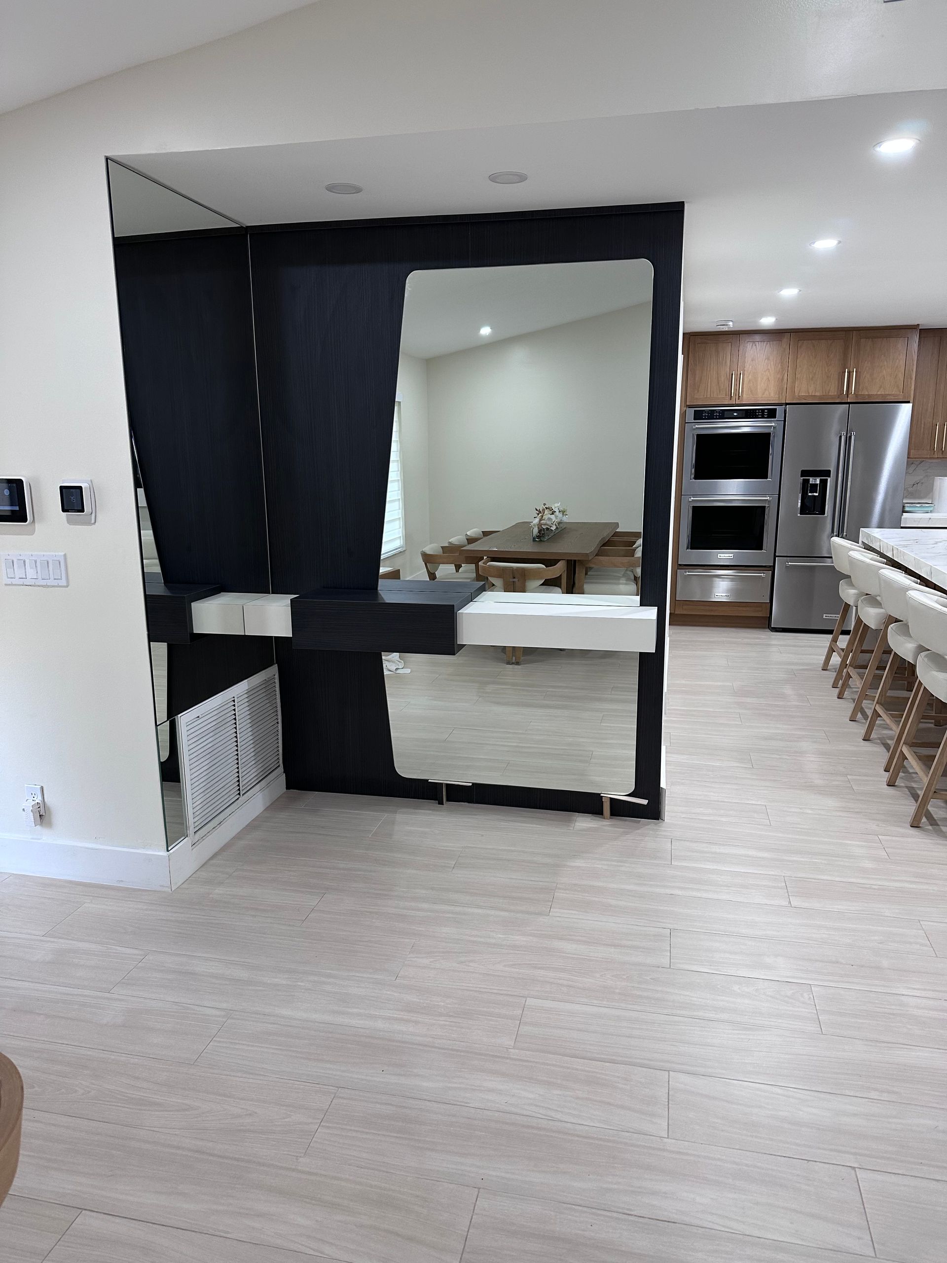 A wide room features a large, rectangular floor-to-ceiling mirror with black trim, reflecting a dining table and kitchen.