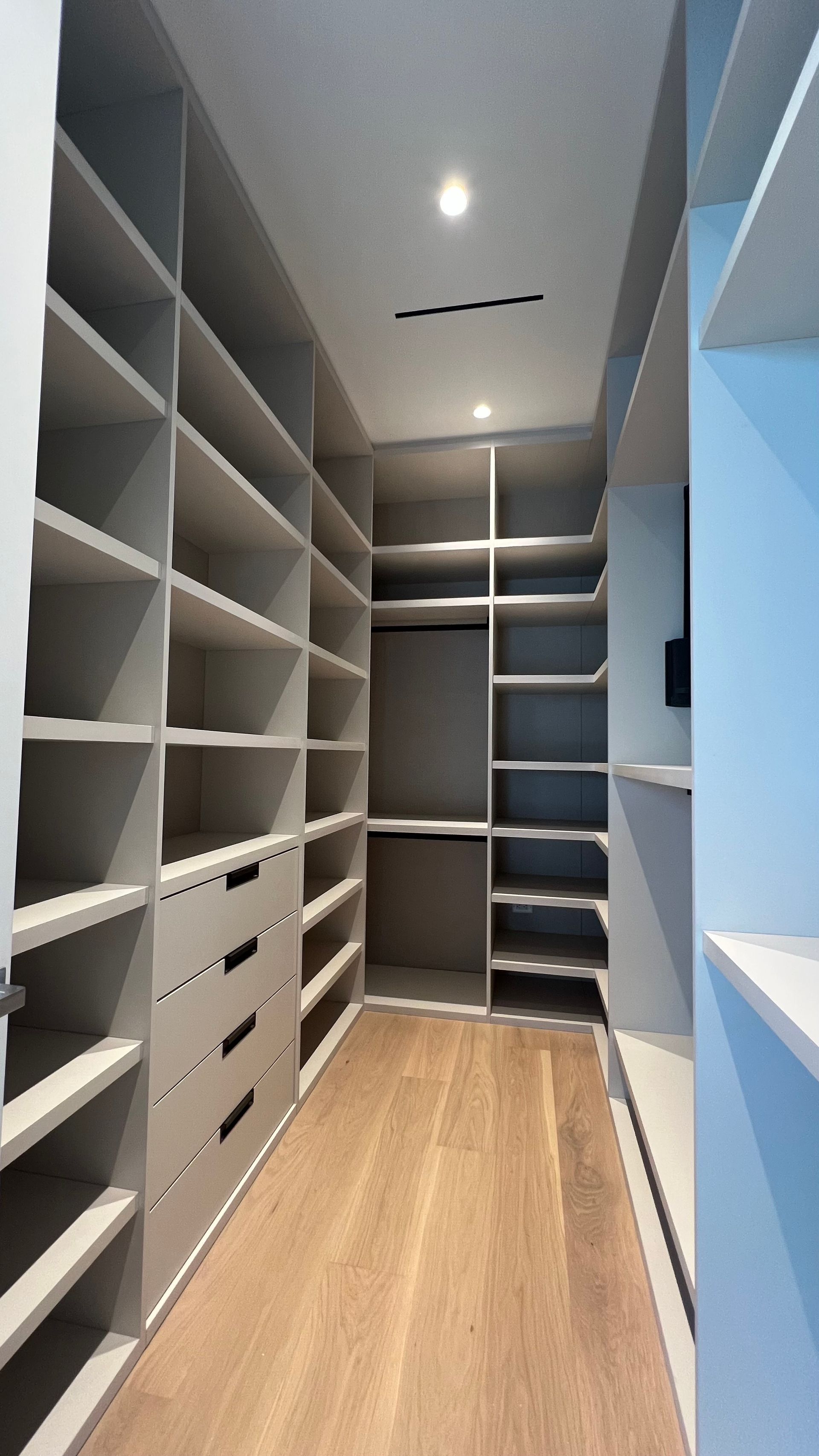 A modern walk-in closet featuring light gray floor-to-ceiling shelving, a built-in drawer unit, and light wood flooring.