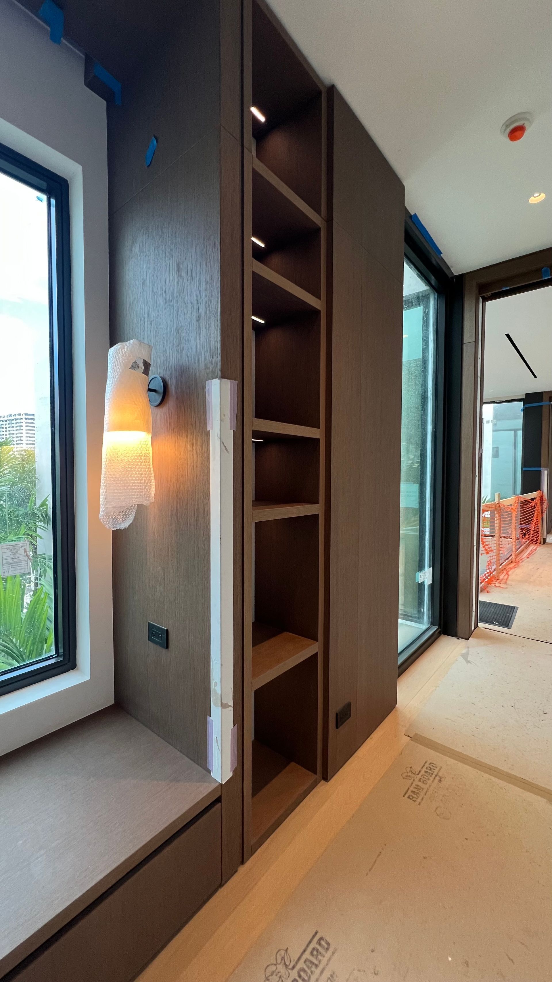 A built-in dark wood shelving unit with integrated lighting sits beside a window with a wall sconce in a room under renovation.