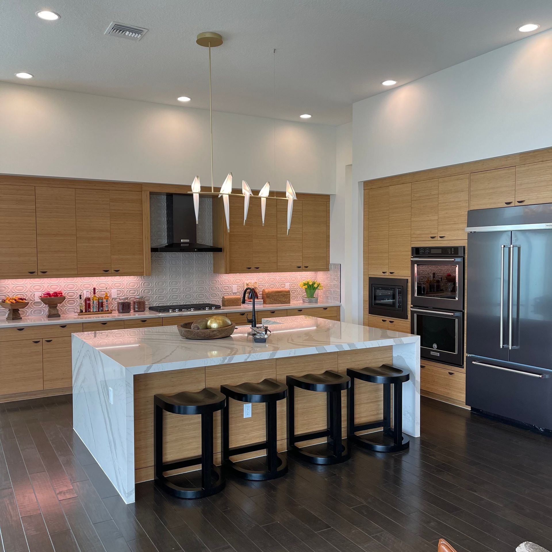 Modern kitchen with light wood cabinetry, white marble island, three black stools, and a unique bird-shaped chandelier.