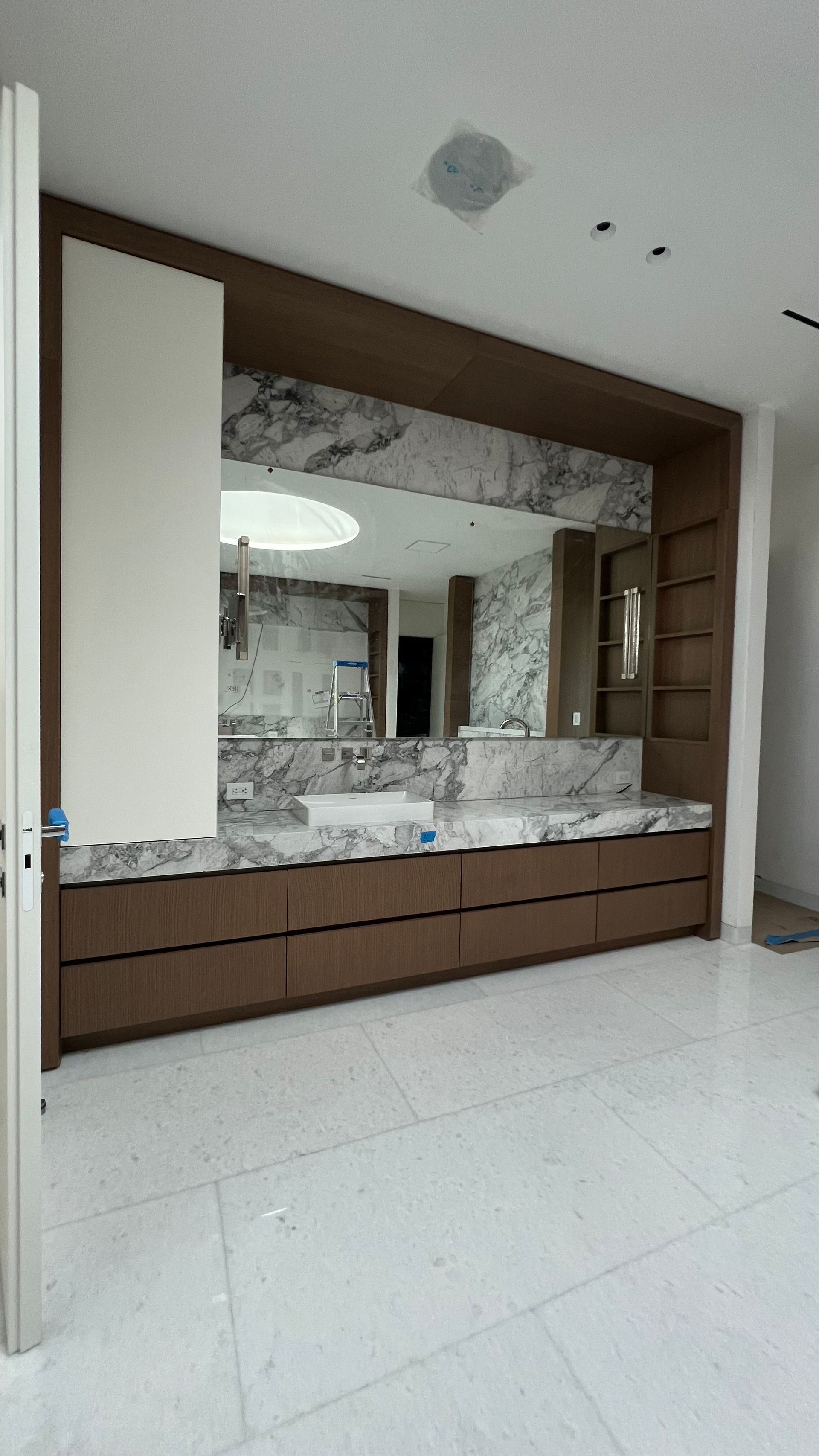 A modern bathroom with a wide wooden vanity, marble countertop and backsplash, and a large wall-to-wall mirror.