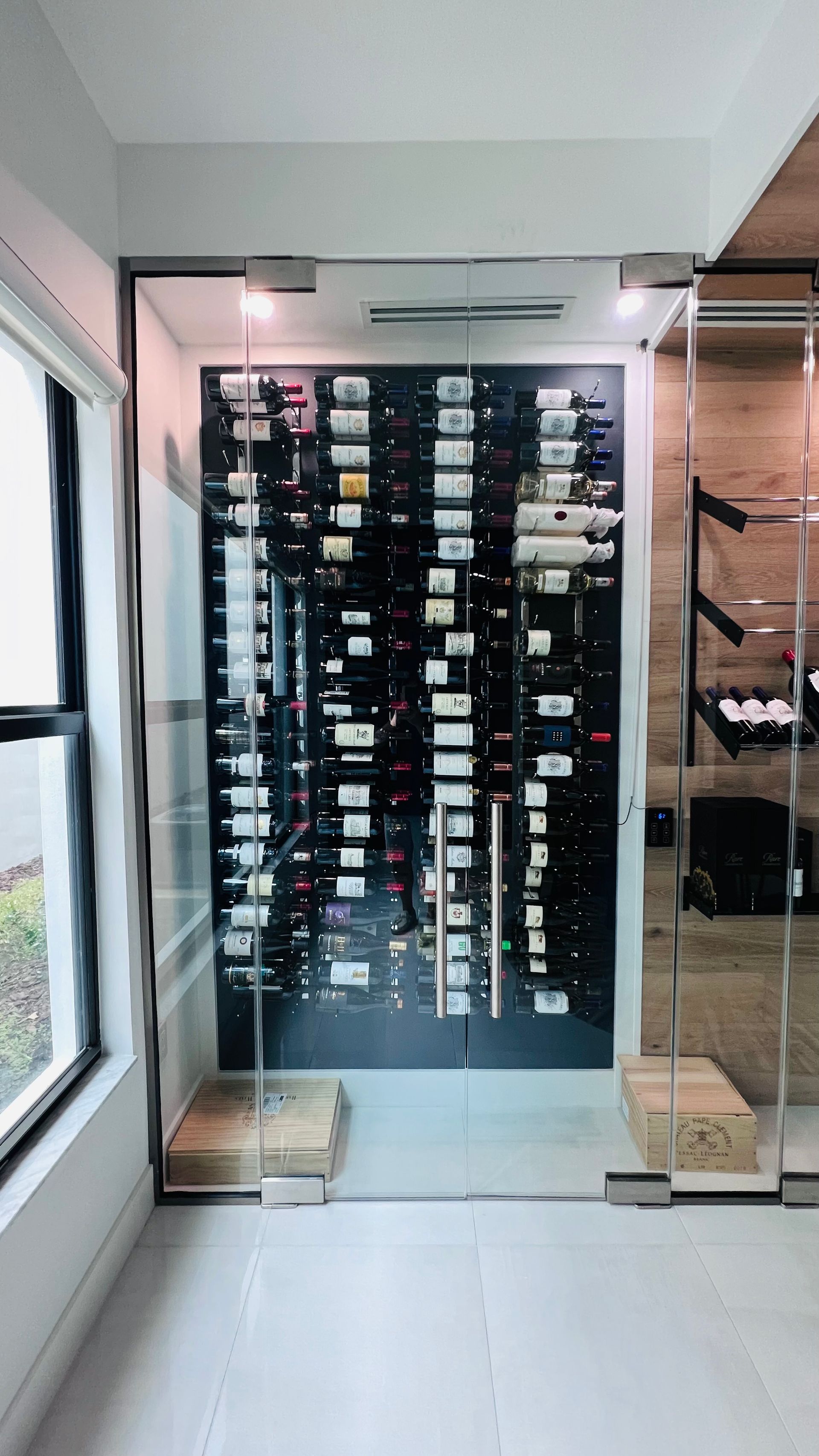 A glass-enclosed wine cellar with vertical metal racks filled with wine bottles against a dark back wall.