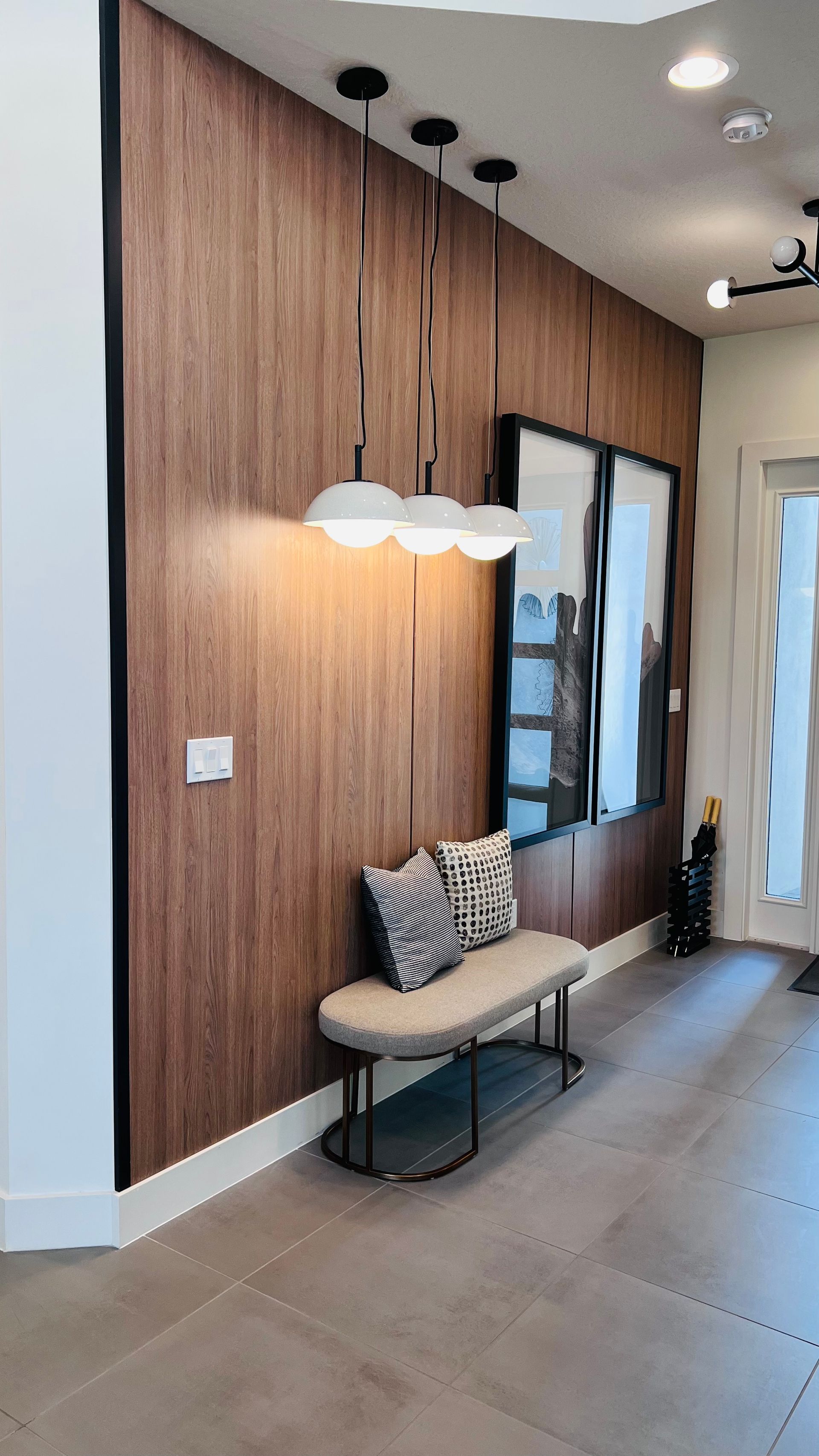 A modern entry area featuring a wood-paneled wall, three globe pendant lights, and a small upholstered bench with pillows.