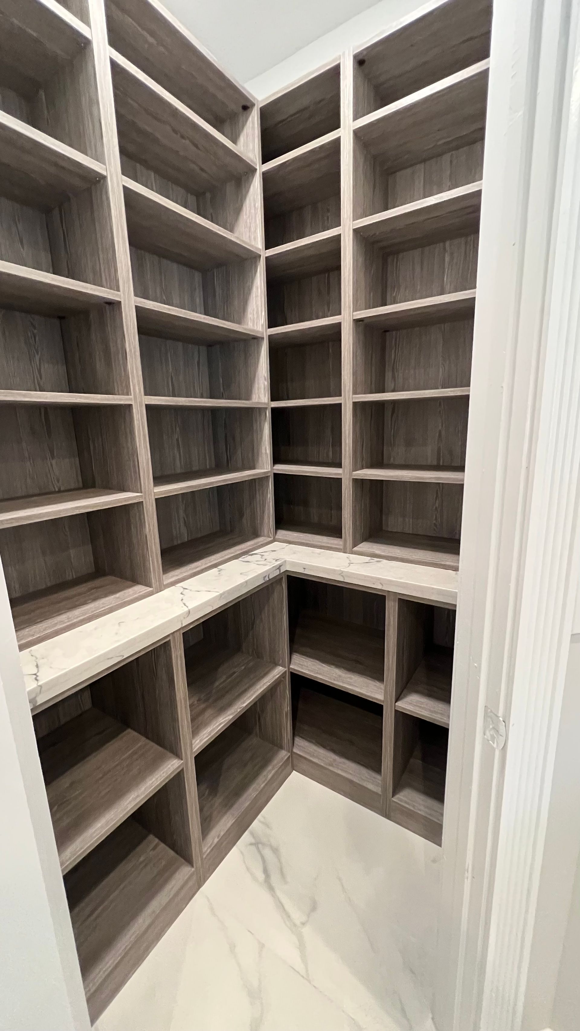 Walk-in pantry with gray wood-grain shelves on white marble-tiled floor.