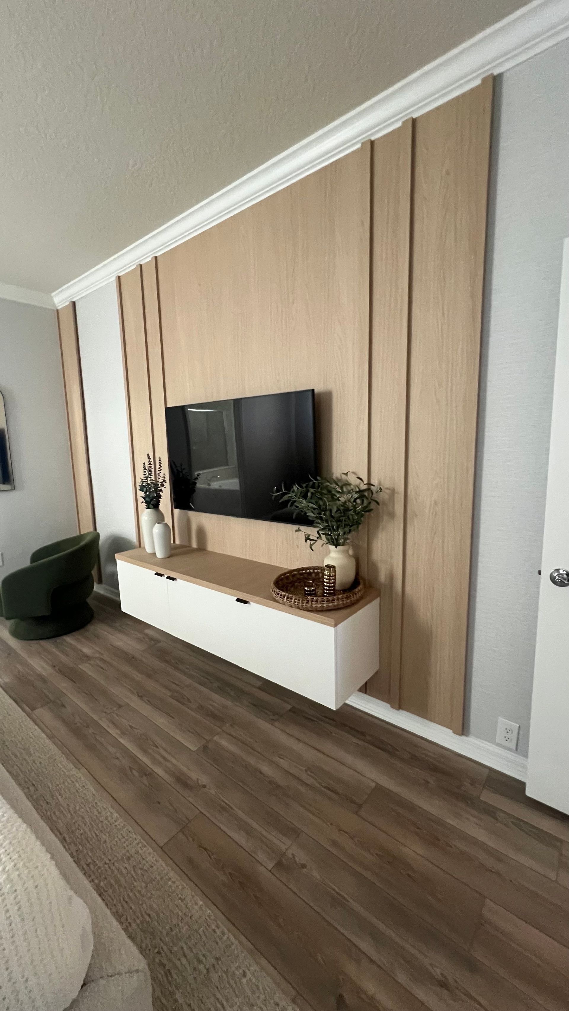 A wall-mounted TV sits above a white floating console on a wooden accent wall, with a small chair to the left.