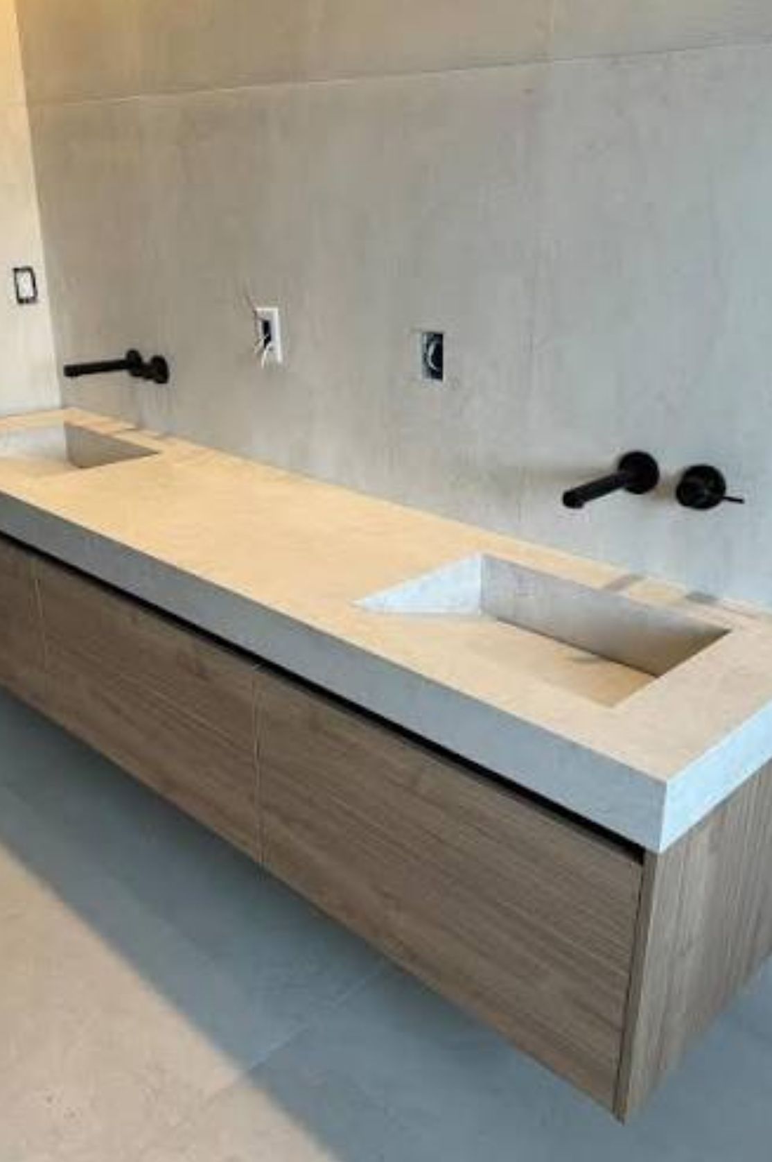 A double vanity with a light stone countertop, wood cabinet, and matte black wall-mounted faucets against a concrete wall.