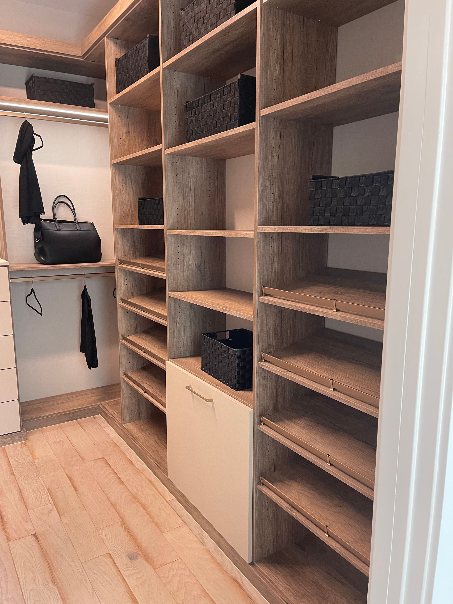 A modern, organized walk-in closet featuring light wood shelving units, a hanging rod, and black storage baskets.
