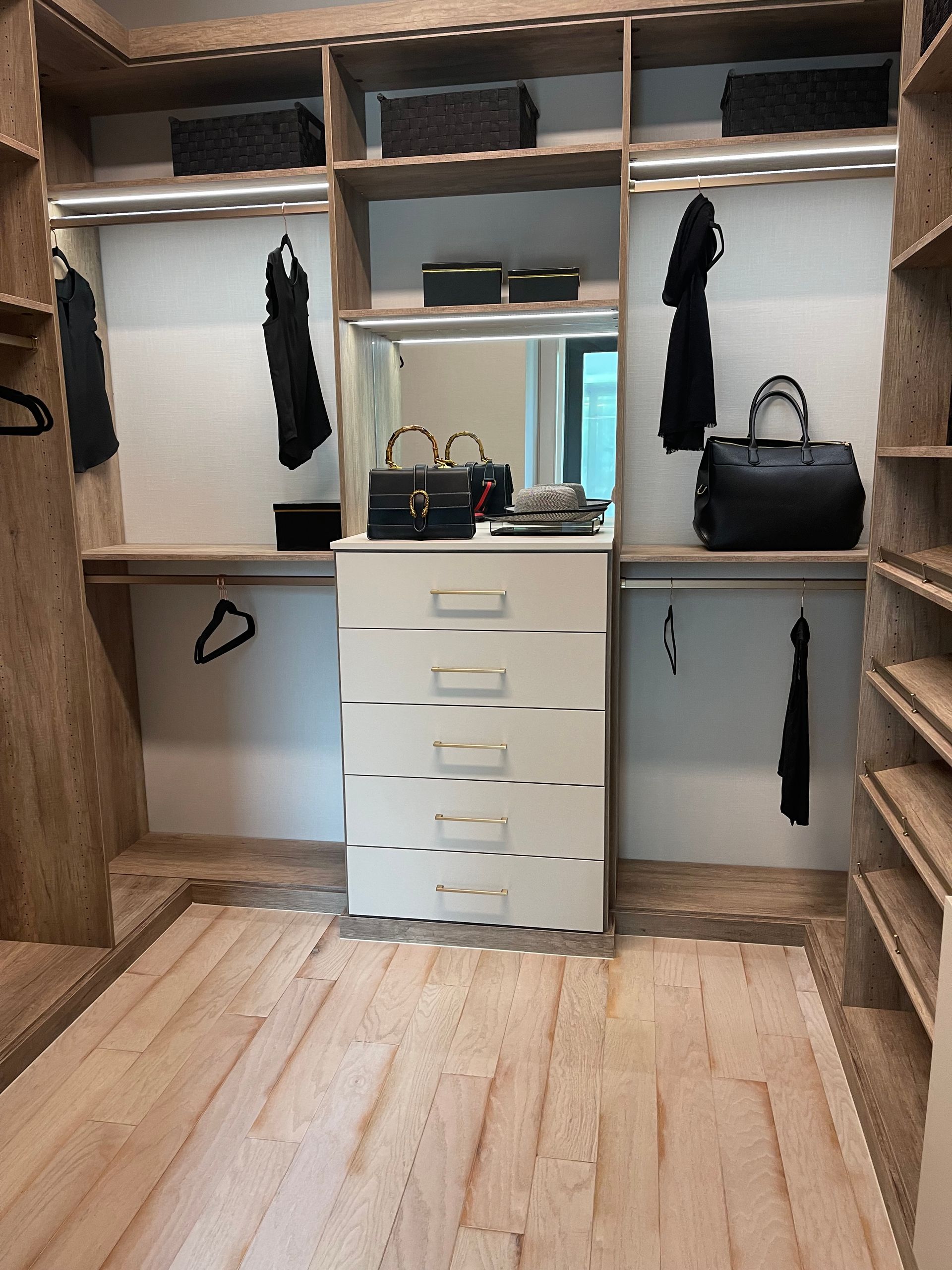 Walk-in closet with wood shelving, hanging clothes, black accessories, and a central white chest of drawers with a mirror.