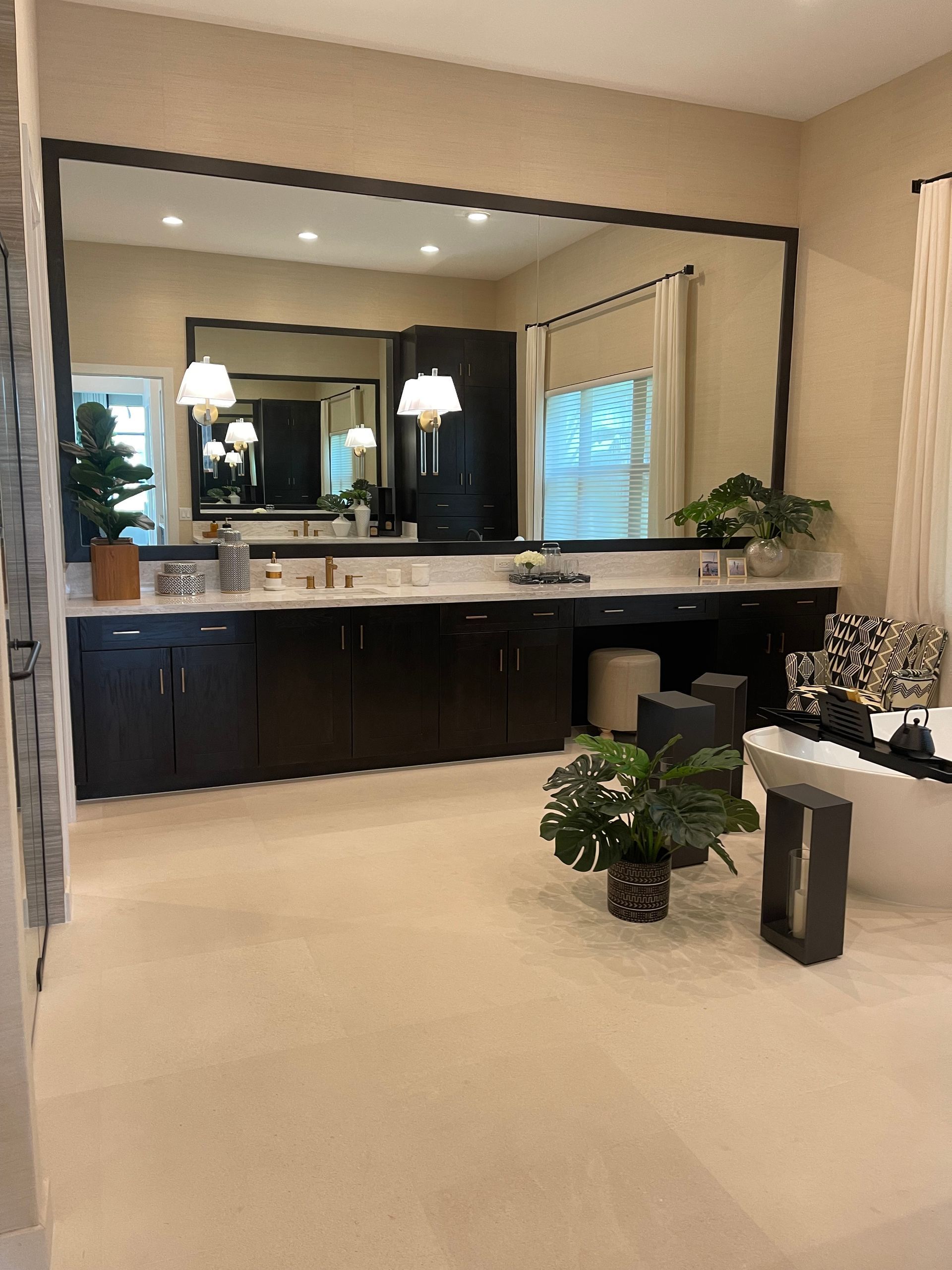 A spacious bathroom with dark wood cabinets, a large mirror, potted plants, and a white soaking tub near a window.