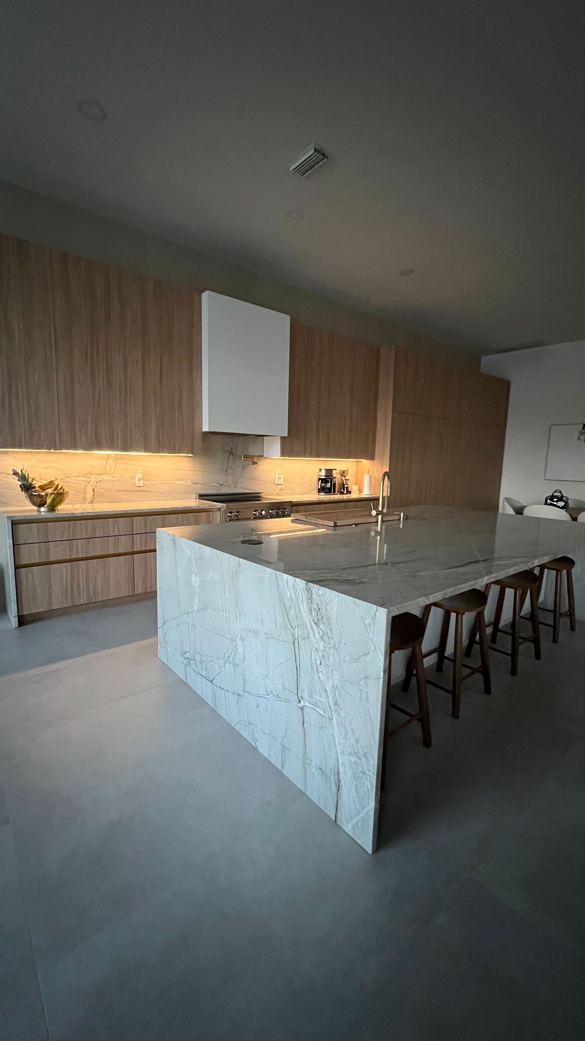 Modern kitchen with wood cabinetry, white marble island, and bar stools over a smooth concrete floor.