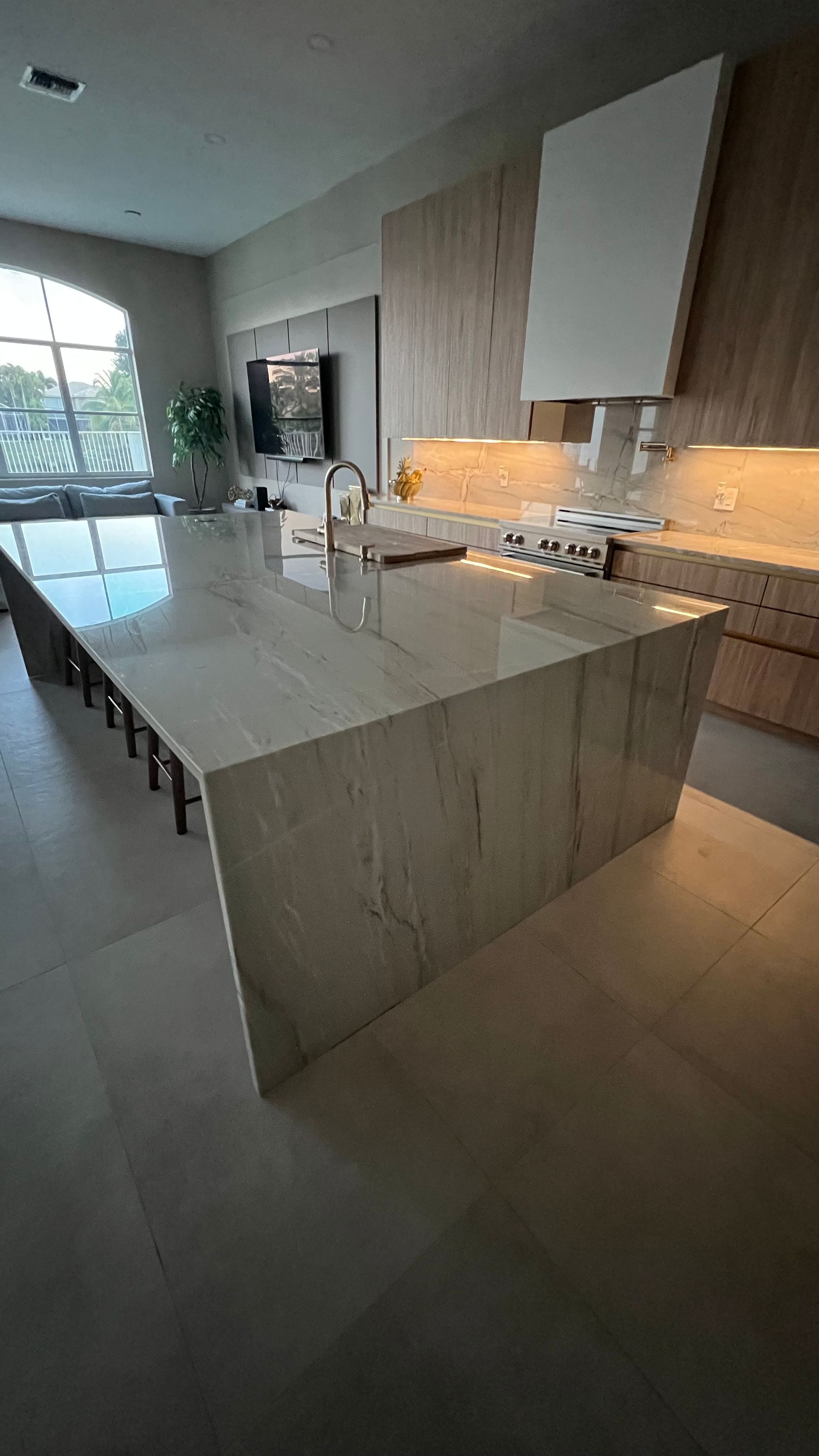 Modern kitchen featuring a large waterfall marble island with bar stools, wood-toned cabinets, and light-colored tile floors.