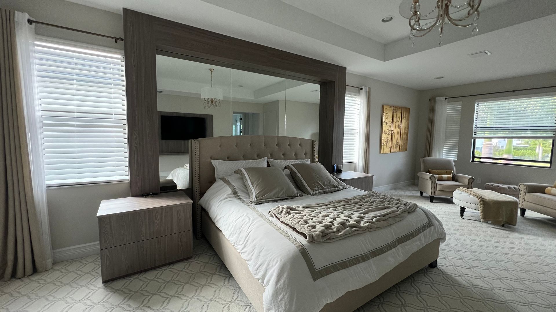 A spacious bedroom featuring a tufted beige bed against a mirror-paneled wall, flanked by light wood furniture.