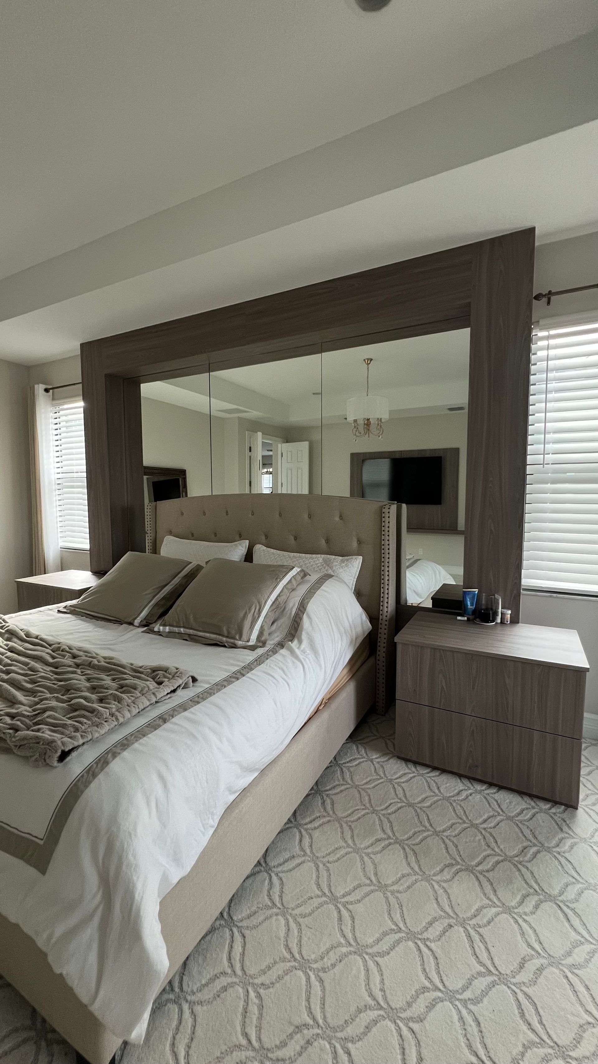 A neutral-toned bedroom features a tufted headboard against a large, floor-to-ceiling mirrored panel framed in wood.