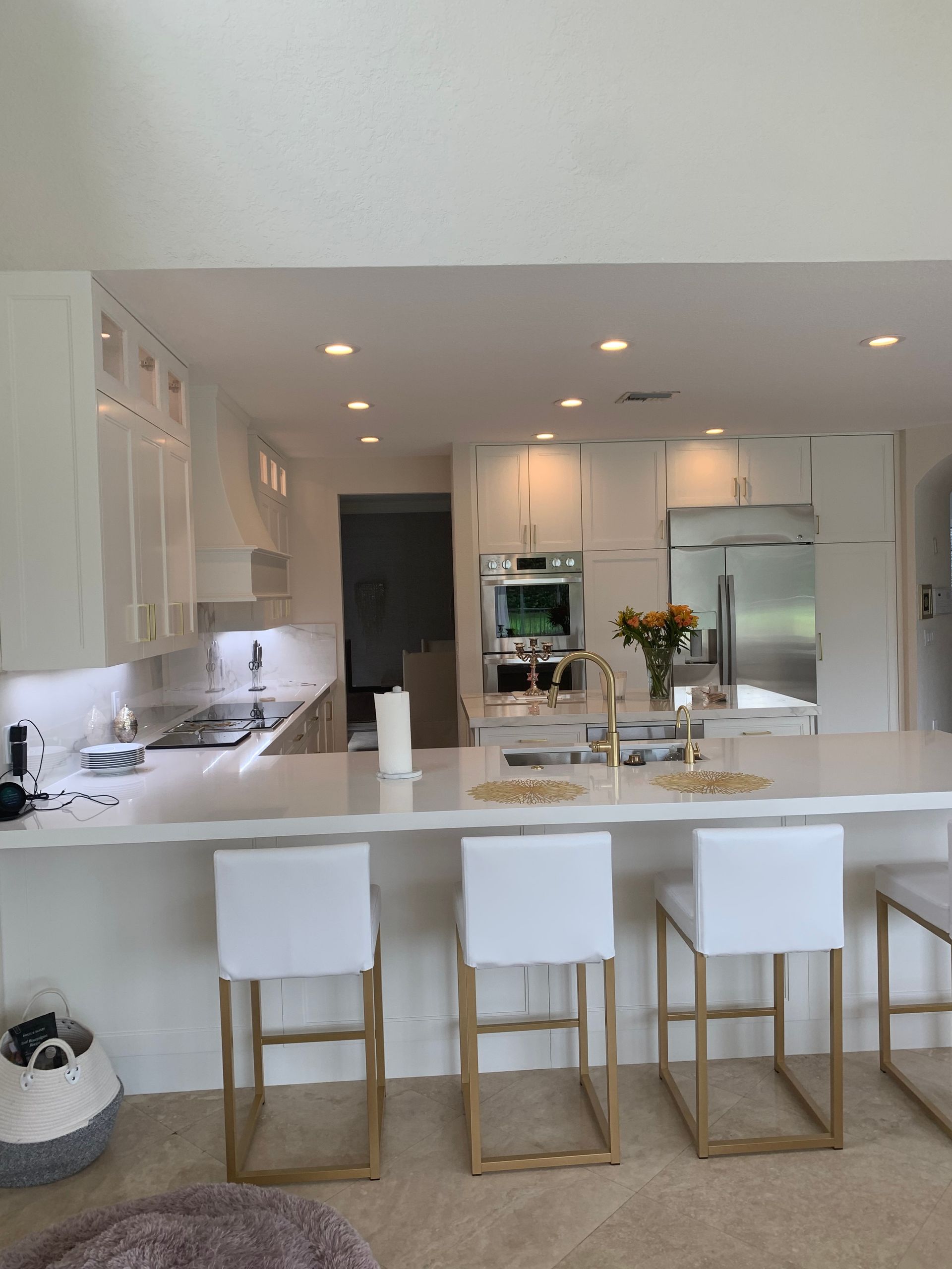 A bright, modern kitchen featuring white cabinets, a large island with three white-and-gold bar stools, and stone counters.