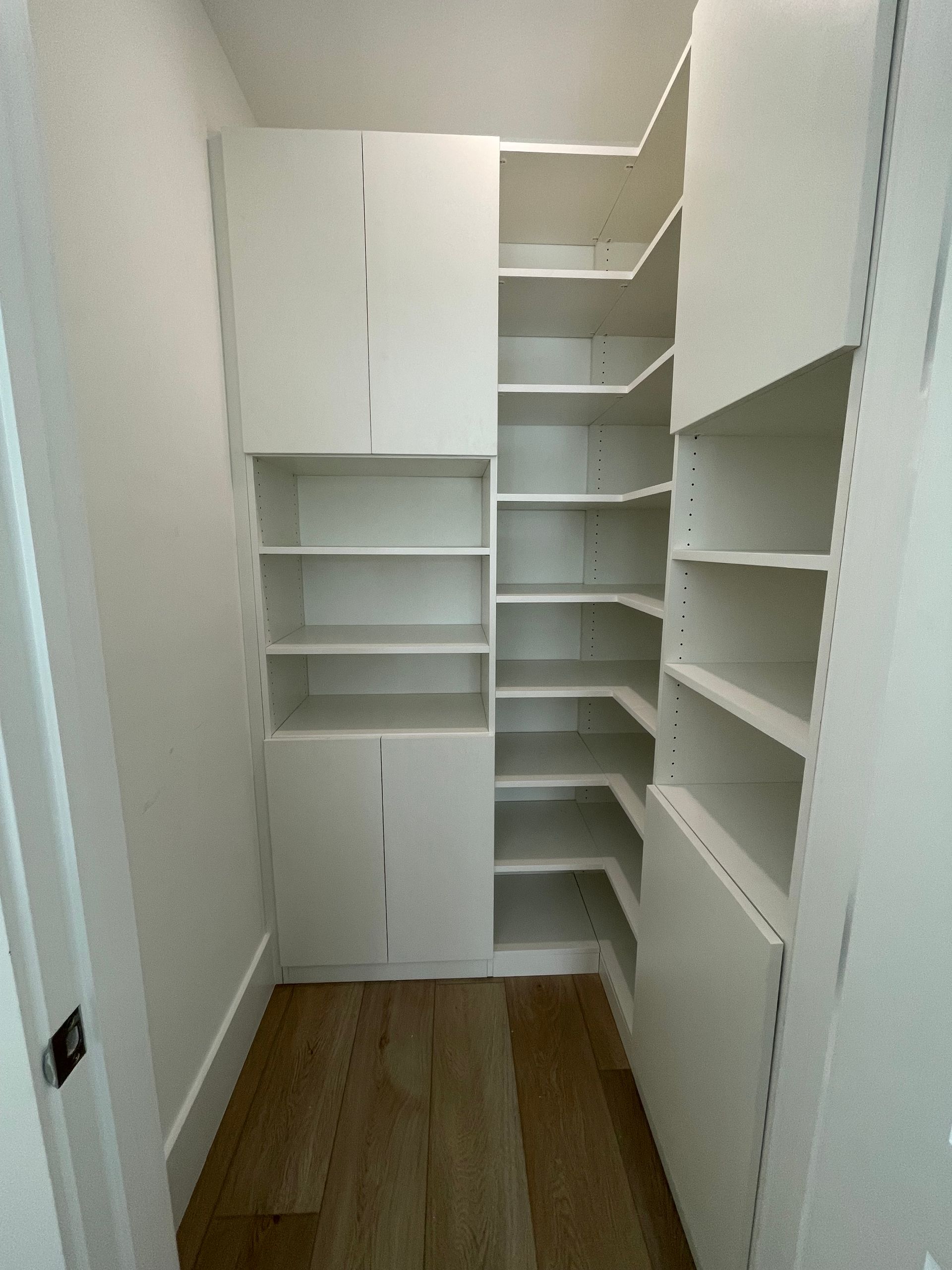 A white, built-in floor-to-ceiling pantry with open shelves and closed cabinet sections, set in a room with wood flooring.